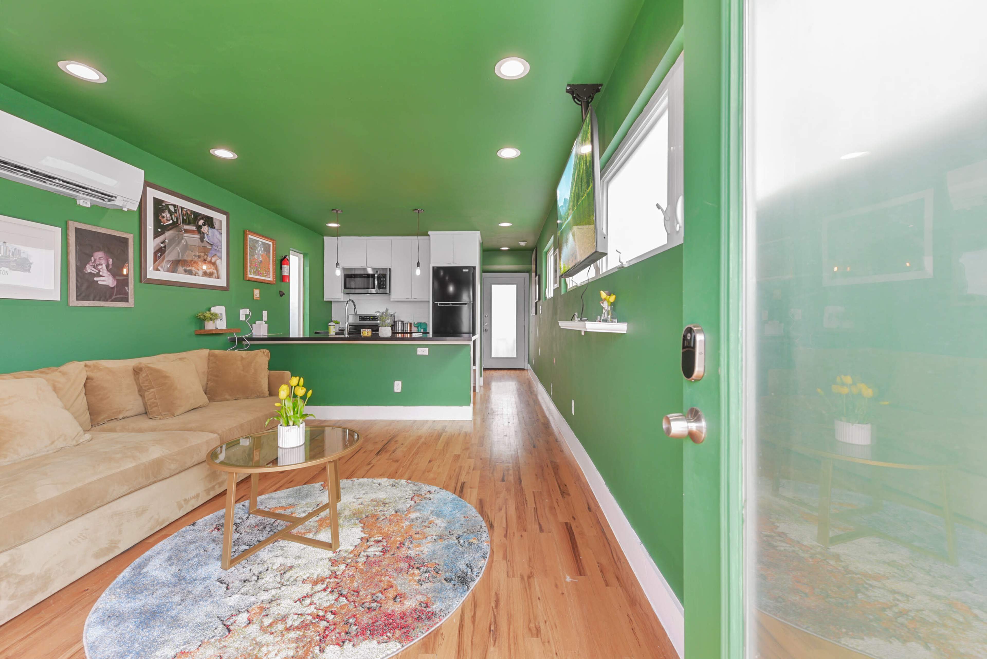 The image shows a cozy living area with green walls, a beige couch, a round rug with a colorful design, and a small kitchen area in the background.