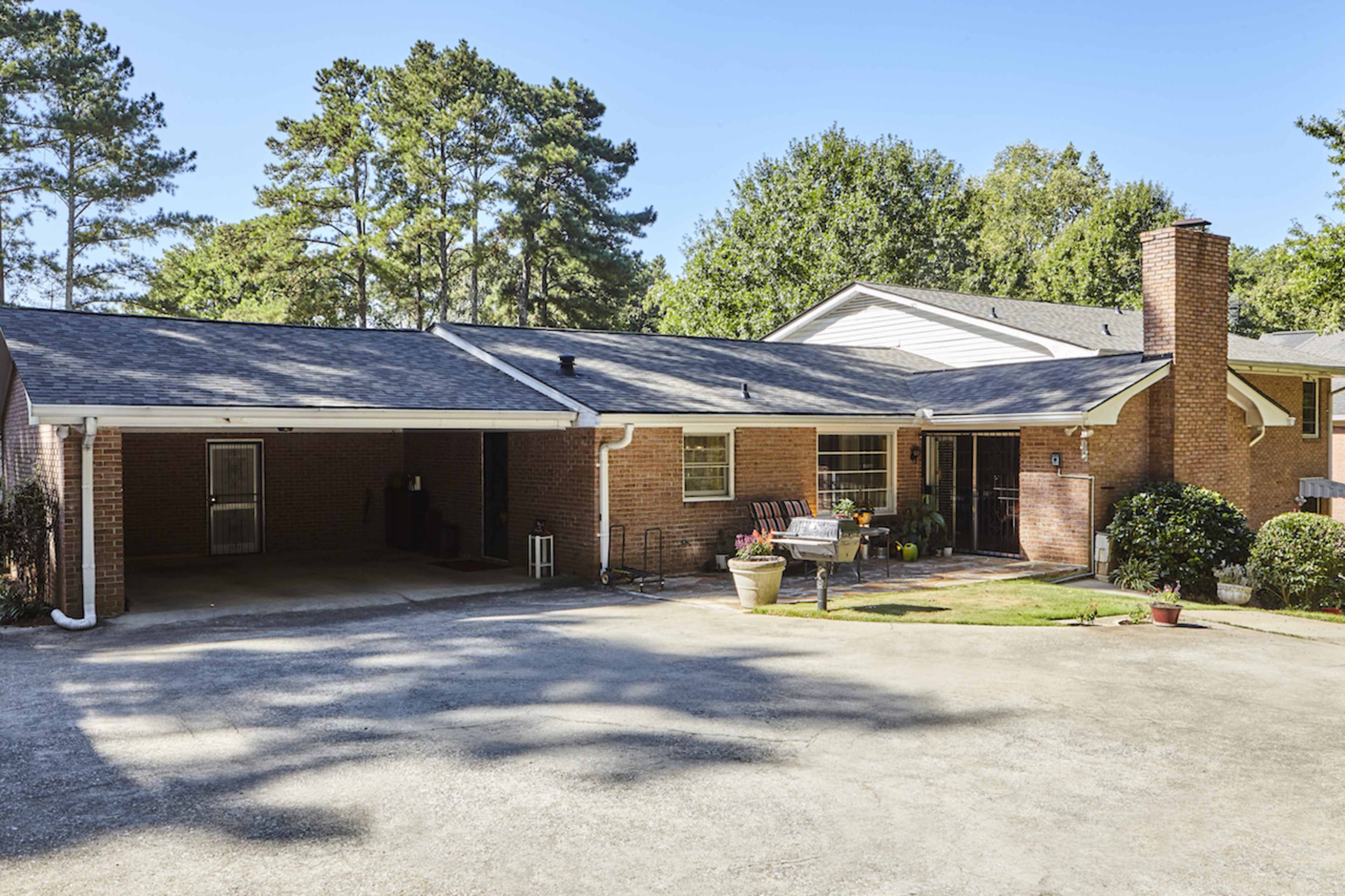A single-story brick house with a carport, surrounded by trees and a gravel driveway.