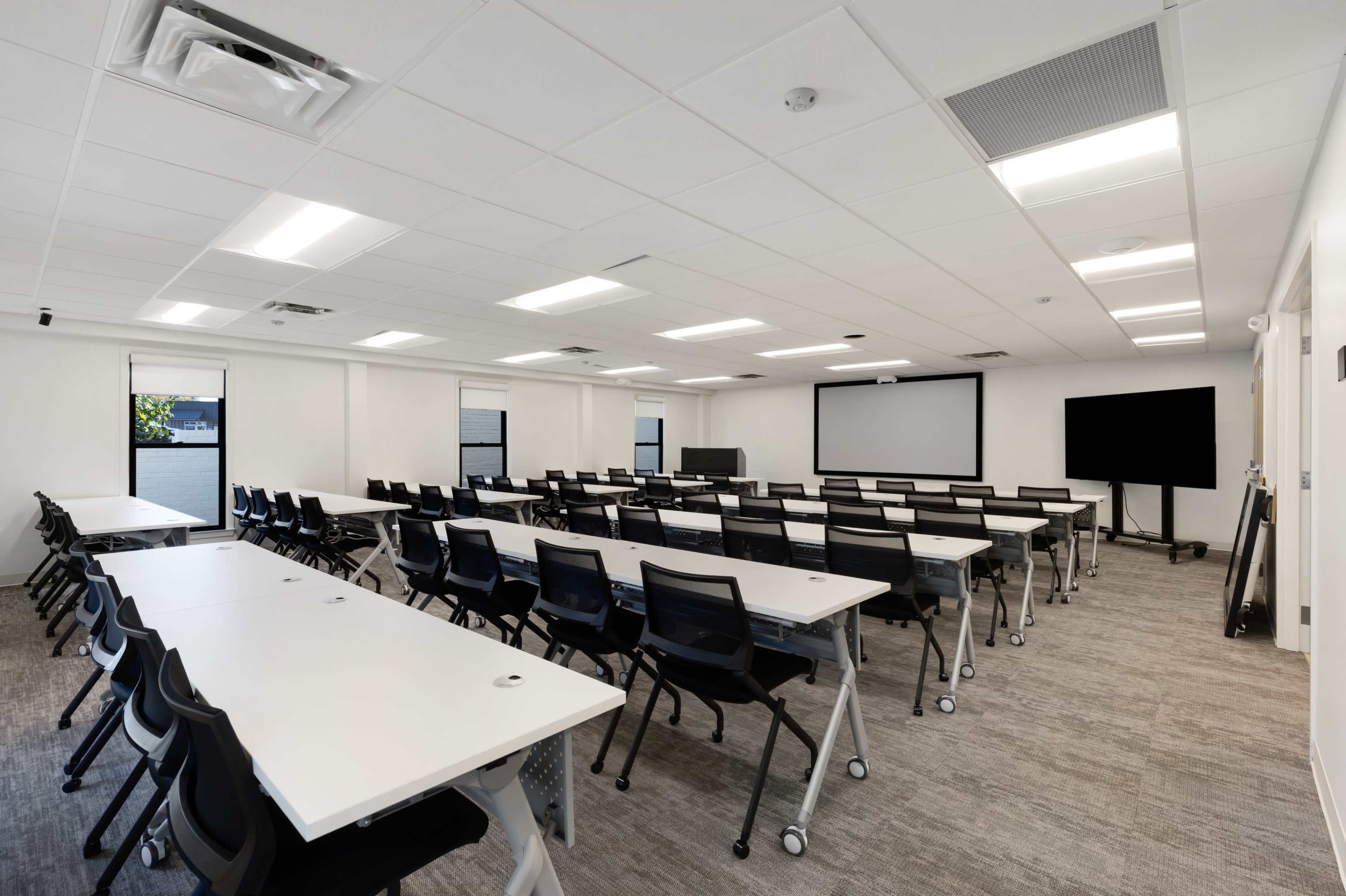 The image shows a modern conference room with several rows of desks and chairs, large windows, and audiovisual equipment on the walls.