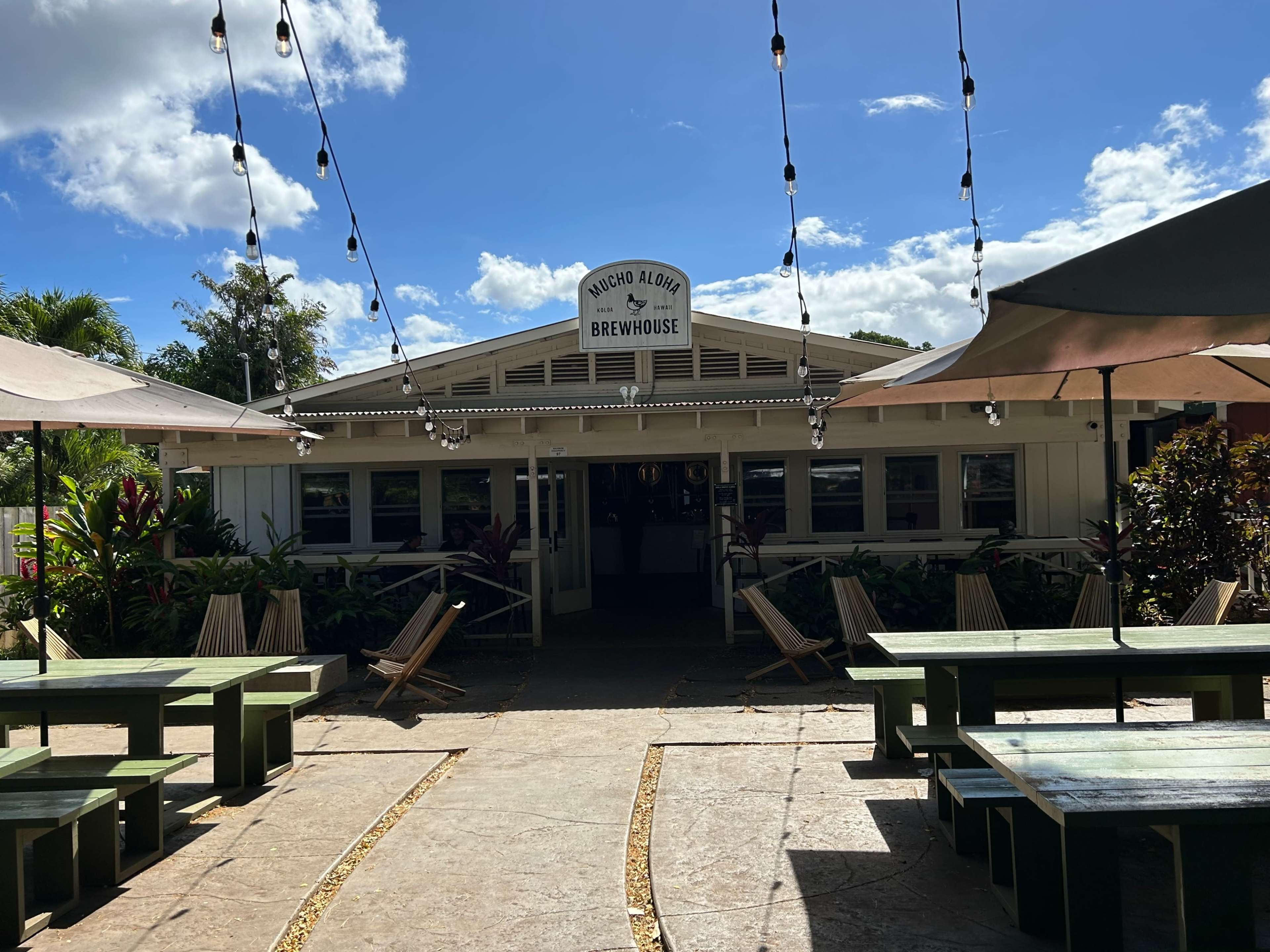 The image shows a brewhouse with a sign that reads "Nacho Aloha," surrounded by outdoor seating and string lights under a partly cloudy sky.