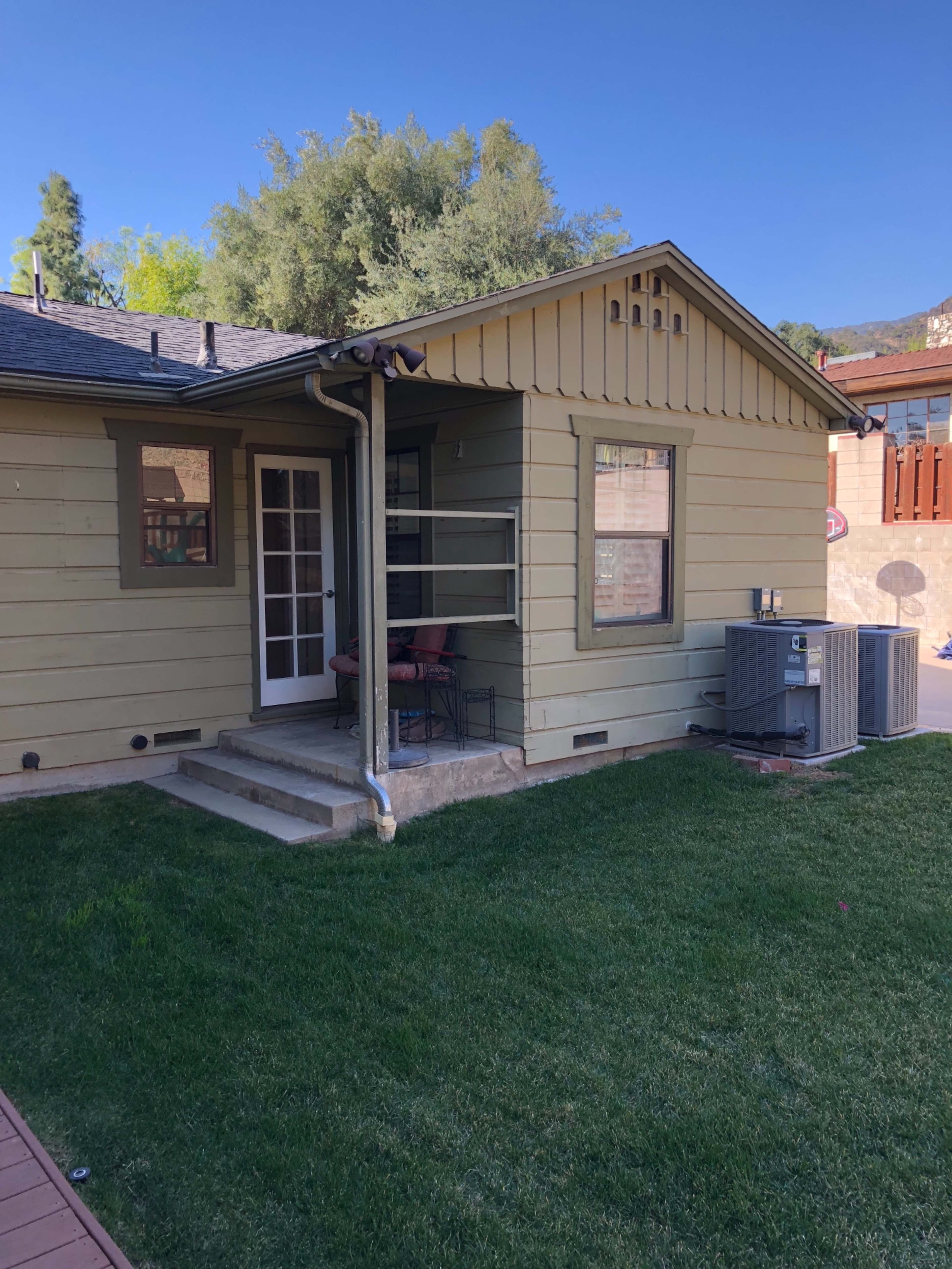 The image shows a small yellow house with a porch and a central air conditioning unit in the yard.