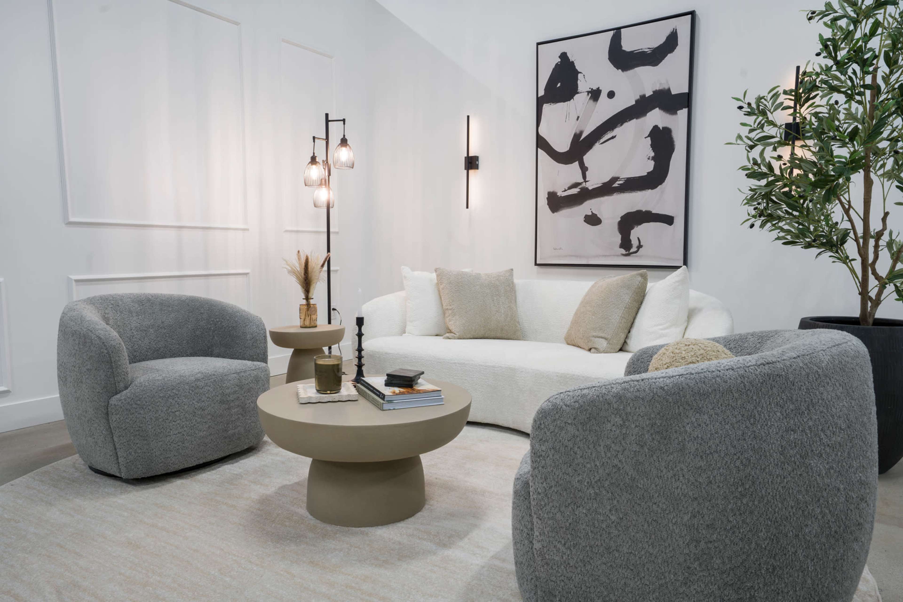 The image shows a modern living room with a white sofa, two gray armchairs, a round coffee table, and a decorative plant.