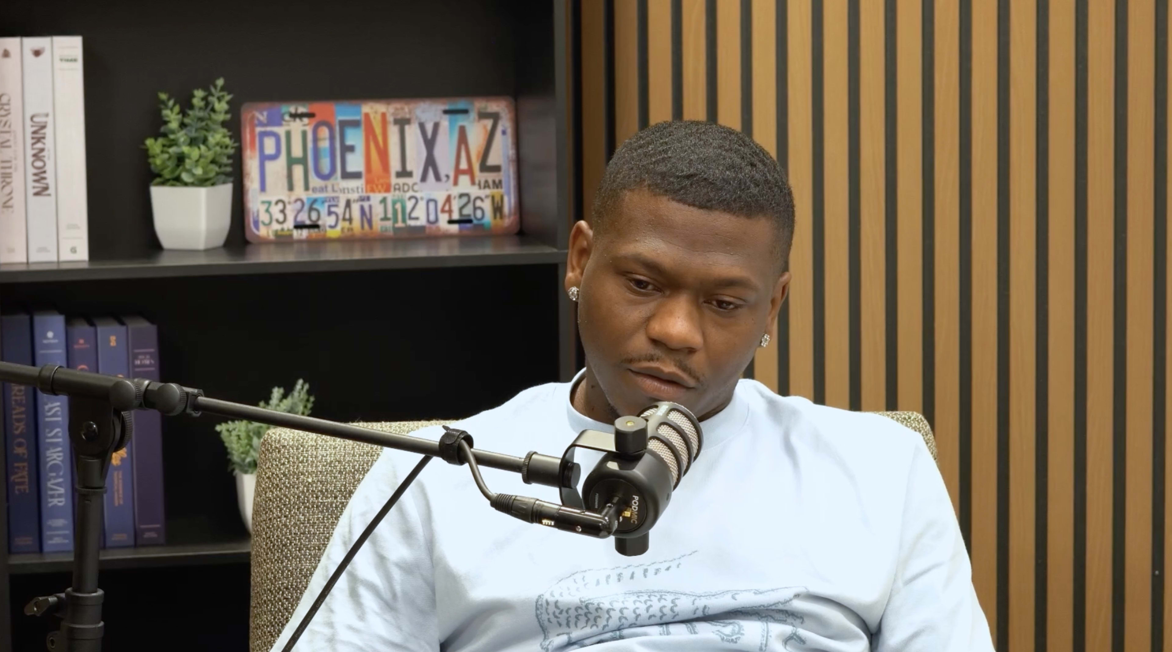 A man sits on a couch in a podcast studio, speaking into a microphone while a decorative background with bookshelves and a license plate is visible.