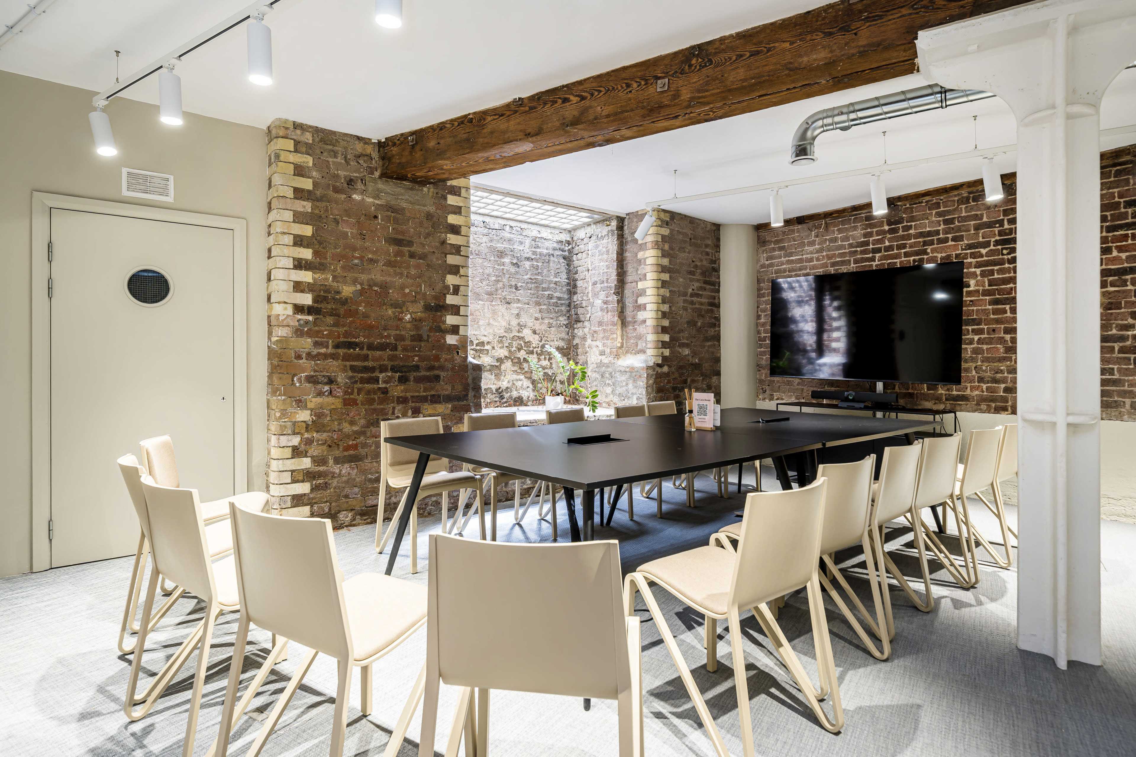 A conference room features a long black table surrounded by beige chairs, with exposed brick walls and a large screen on one side.