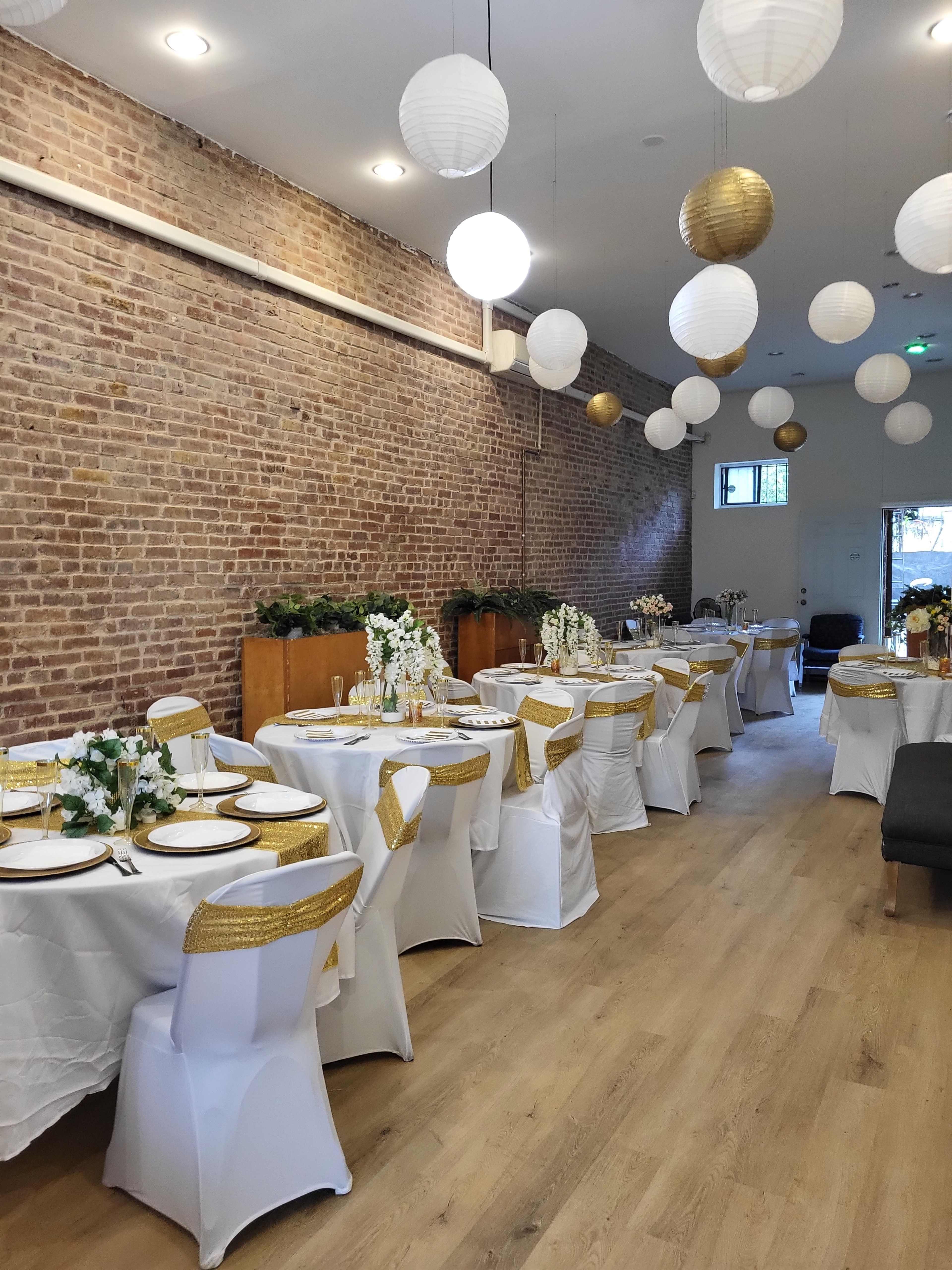 The image shows a decorated dining hall with white tables covered in tablecloths adorned with gold accents, set with floral centerpieces under hanging paper lanterns.