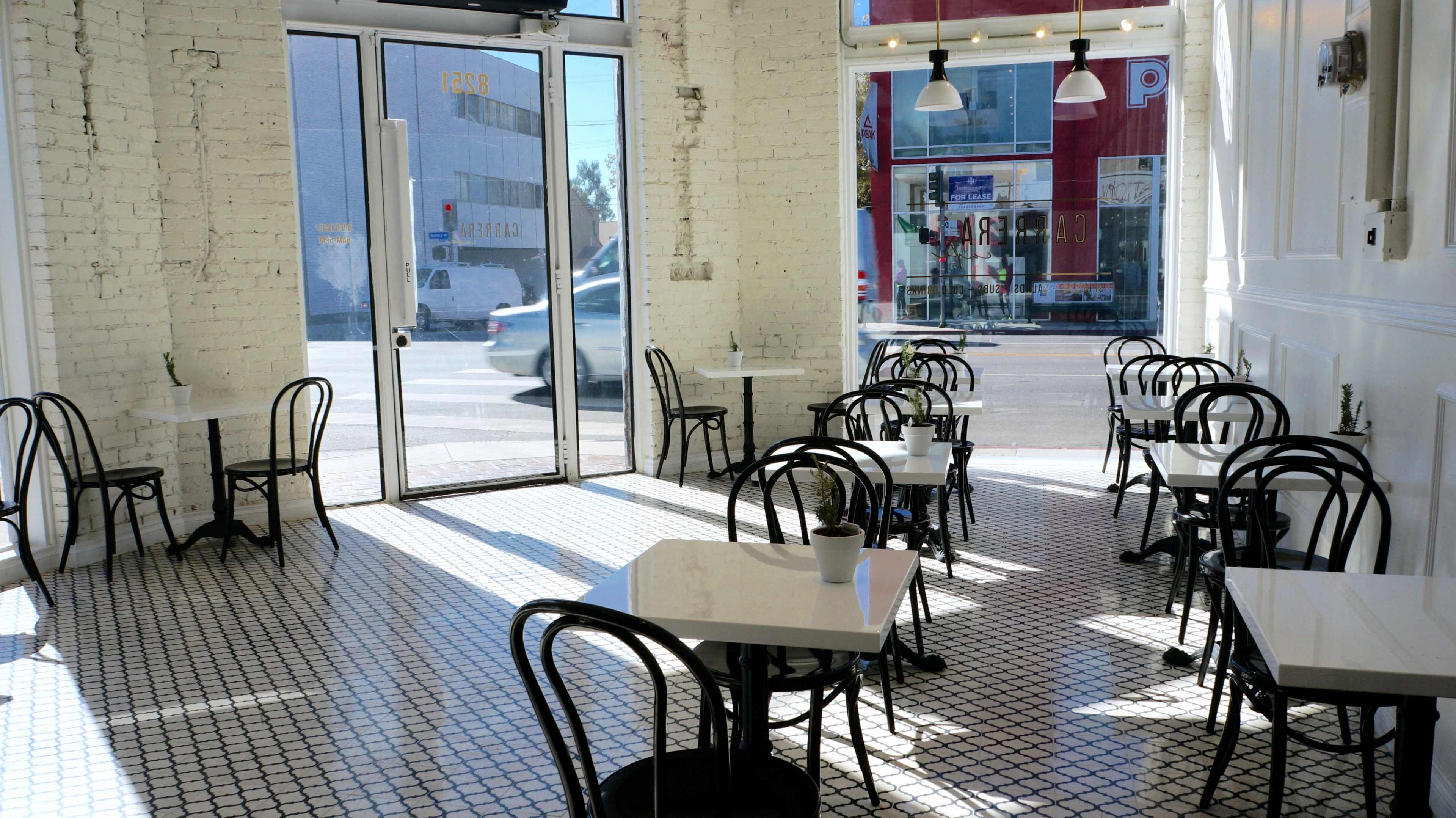 A bright, modern café features black bistro chairs and white tables arranged on a tiled floor with large windows letting in natural light.