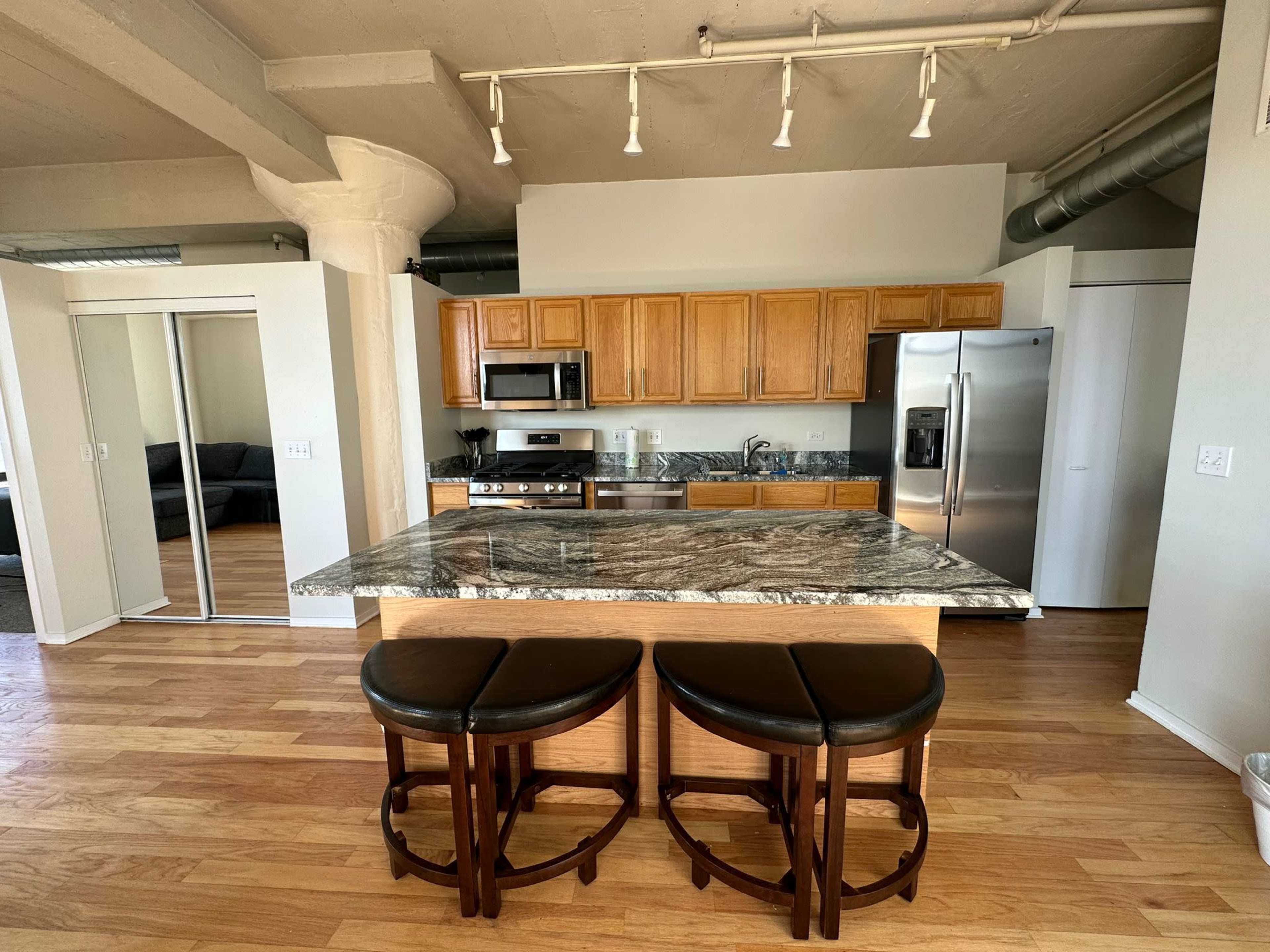 A modern kitchen with wooden cabinets, stainless steel appliances, and a large granite-topped island with two black bar stools.