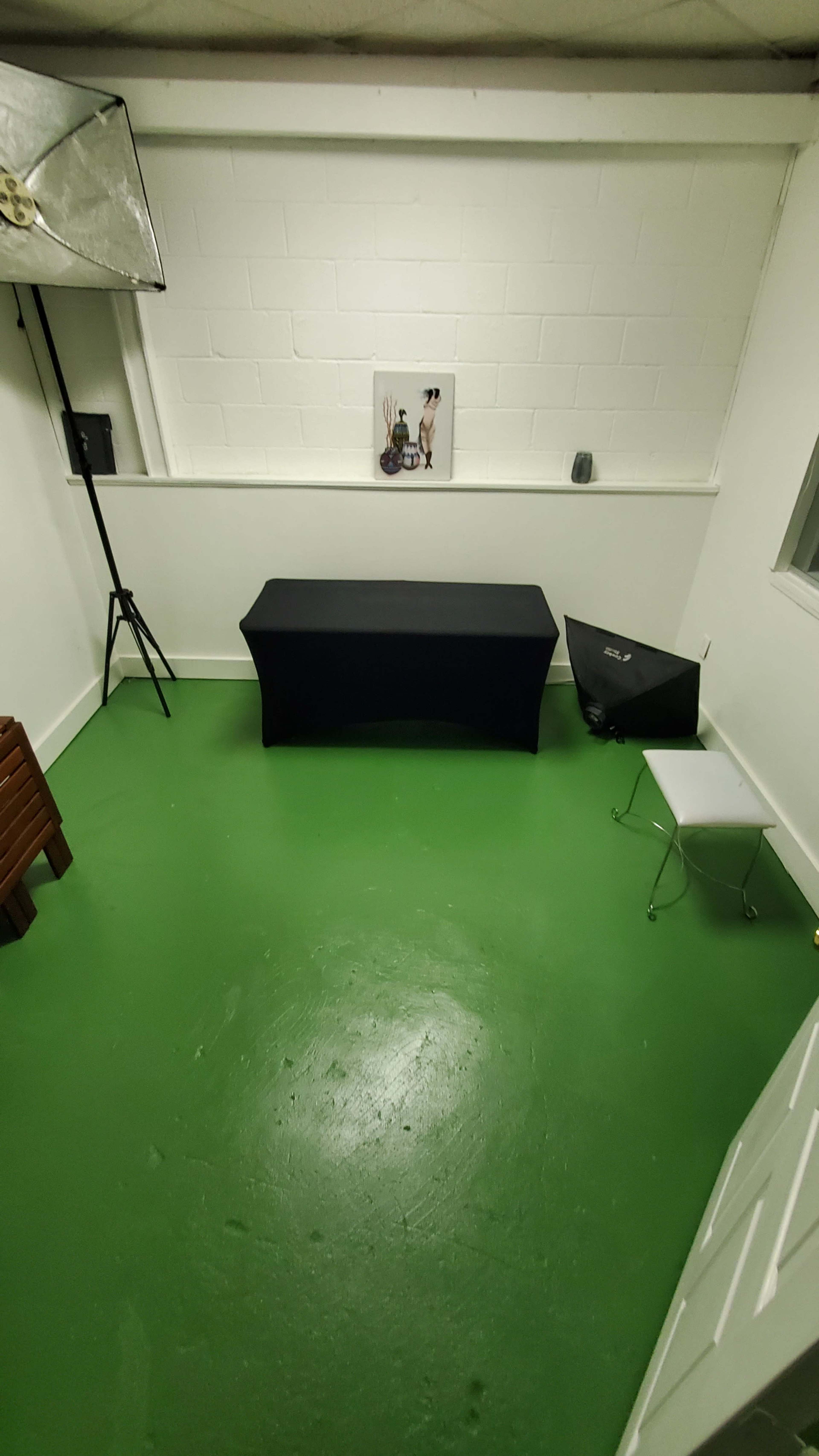 The image shows a small, empty room with green flooring, a black table, a chair, and a wall-mounted picture.