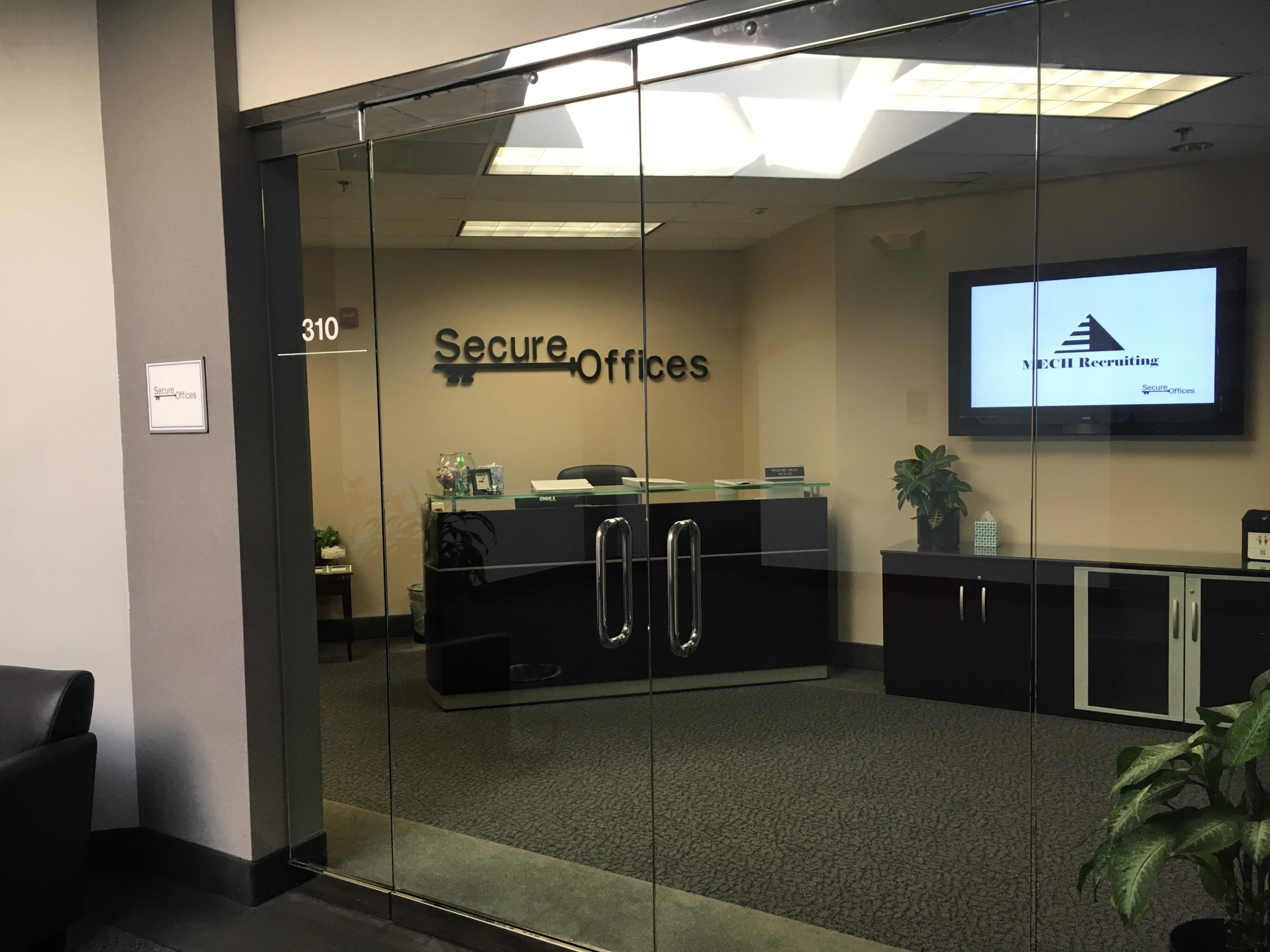 A glass entrance to an office labeled "Secure Offices," featuring a reception desk and a television display.