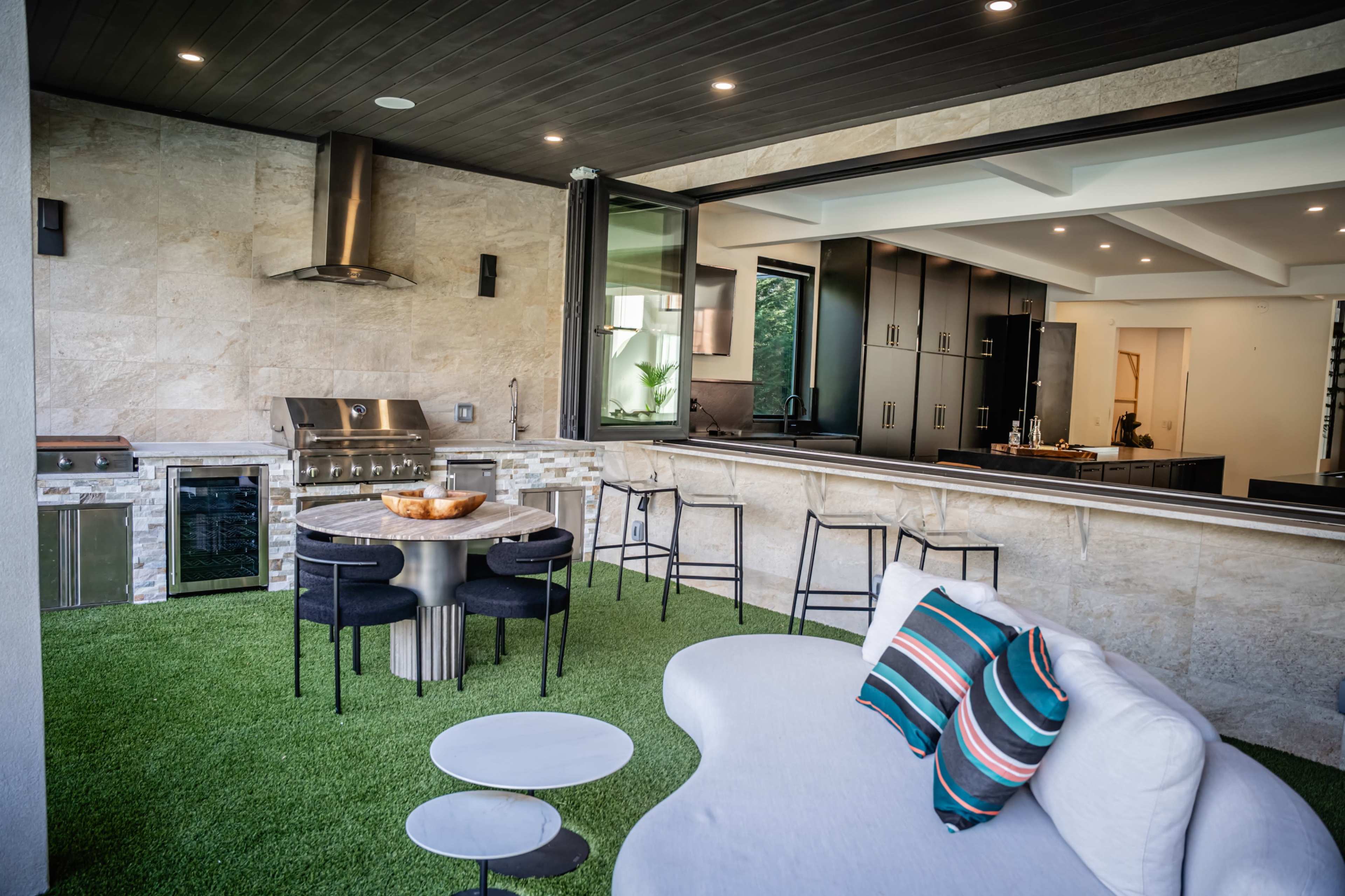 The image shows a modern outdoor kitchen and dining area with a built-in grill, sleek cabinetry, and a grass-like floor covering.