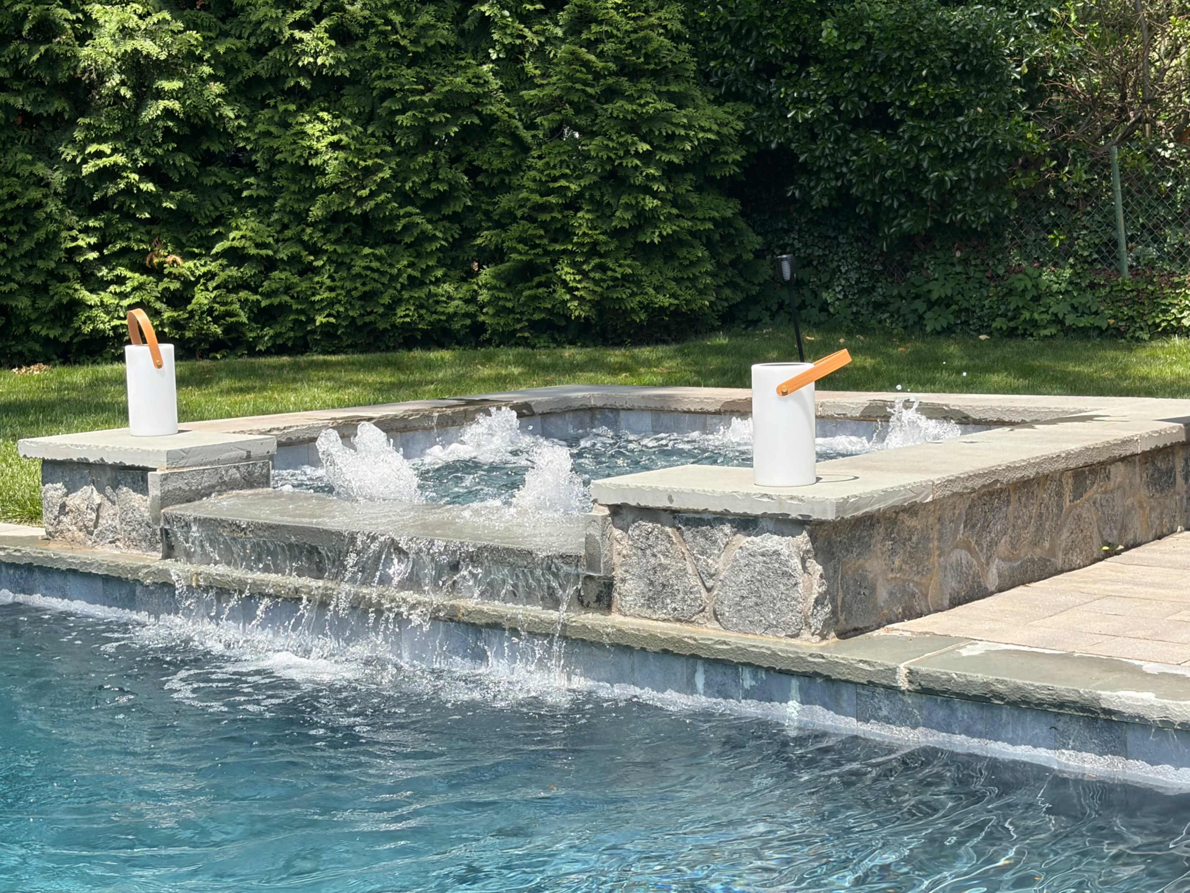 A spa with a waterfall feature is located adjacent to a swimming pool, surrounded by green landscaping.