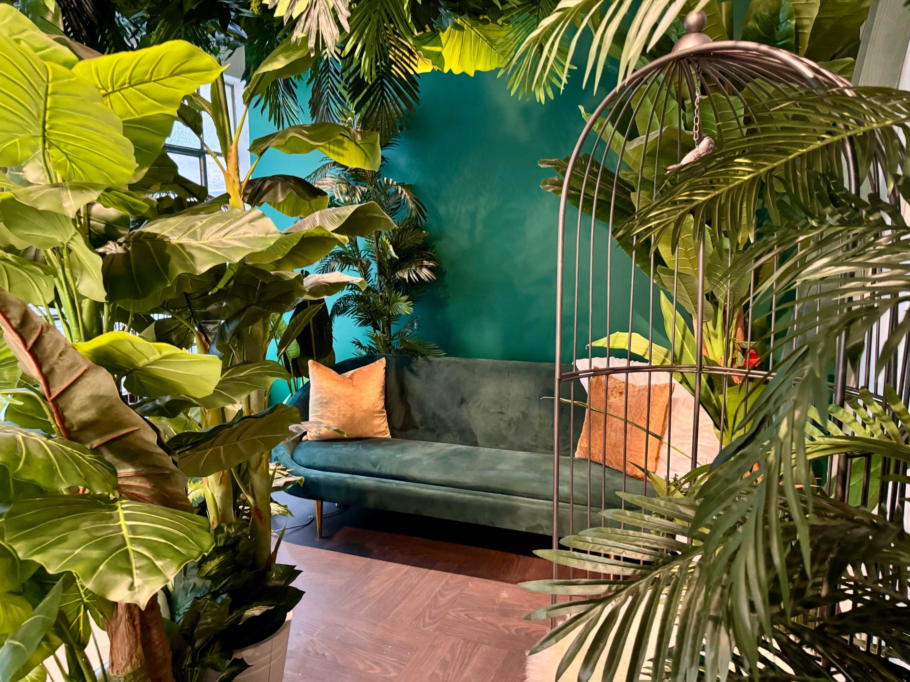 The image features a green velvet sofa positioned against a teal wall, surrounded by various indoor plants and decorative pillows.