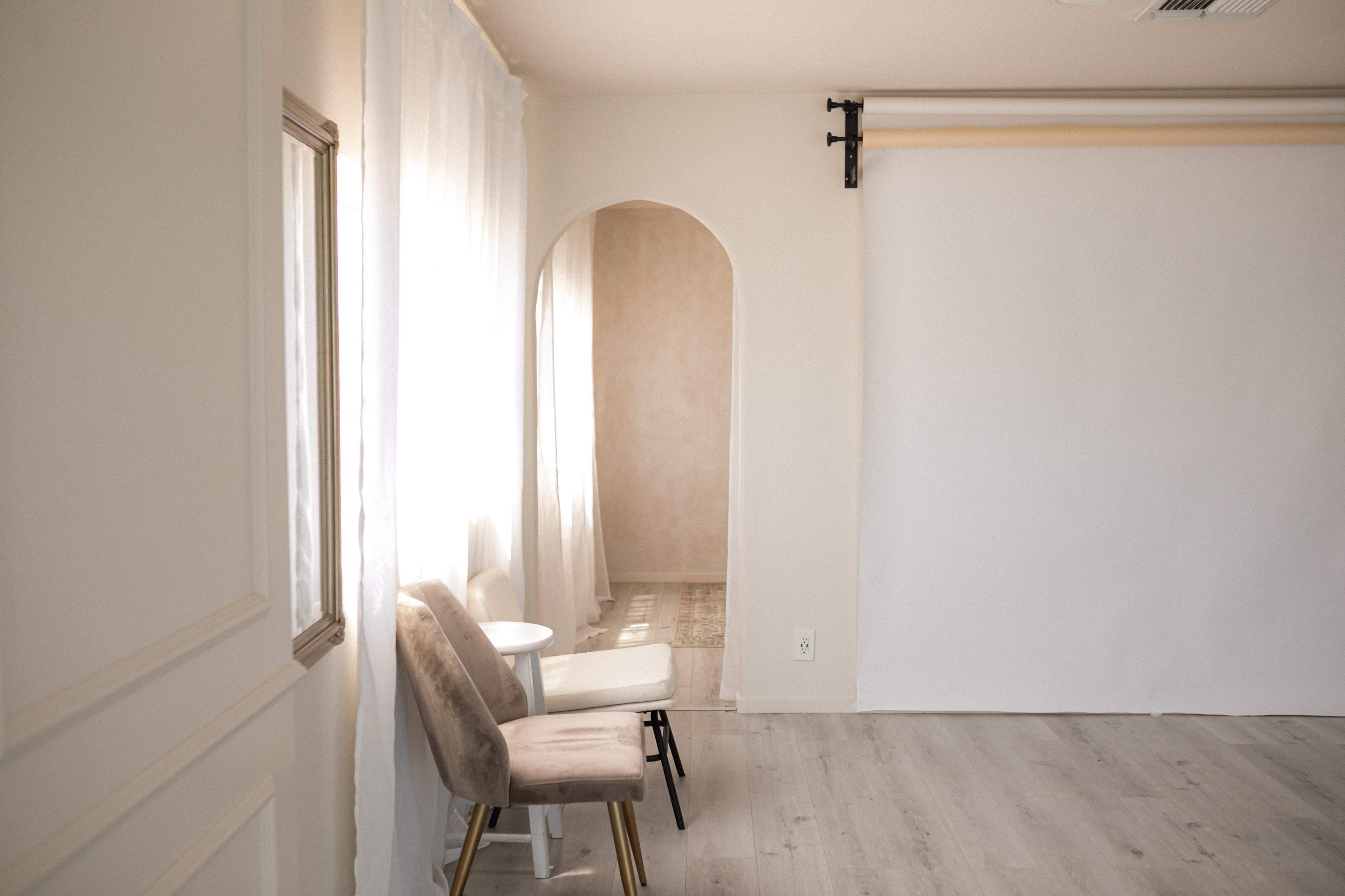 A minimalistic room featuring two chairs beside a small table, with an archway leading to a softly lit area and a blank backdrop on the wall.