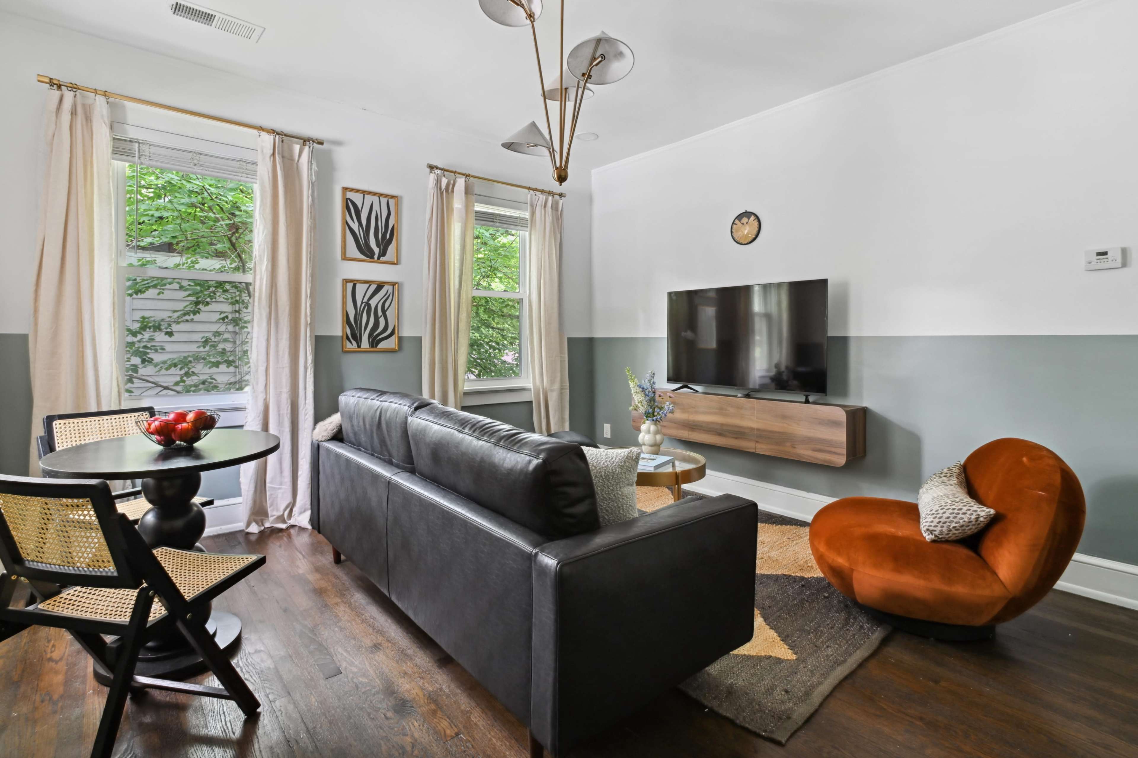 A living room features a black leather sofa, an orange chair, a wall-mounted television, and a small dining table with a bowl of apples.