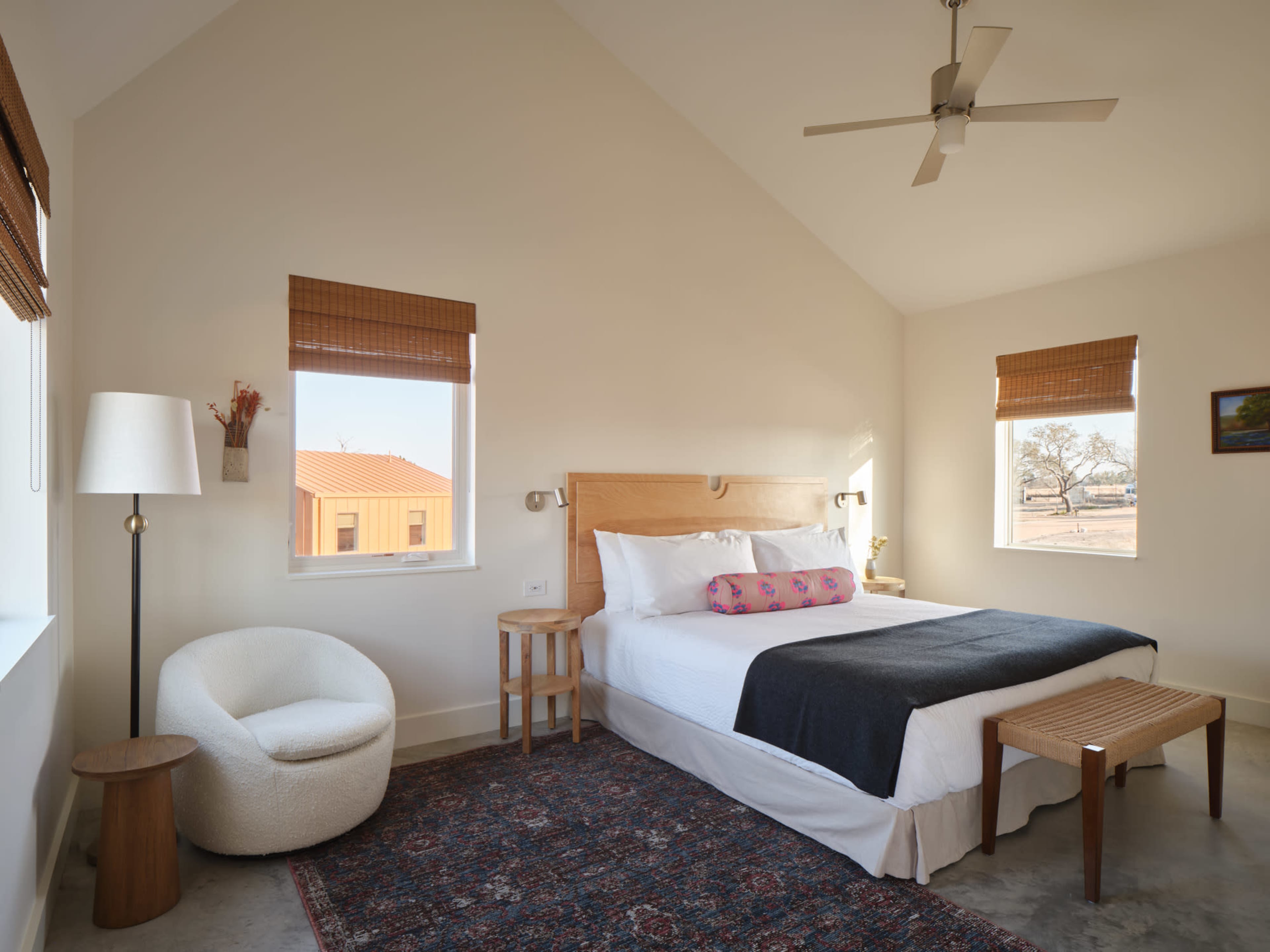 The image shows a minimalist bedroom with a large bed, wicker elements, and bright natural light coming through two windows.