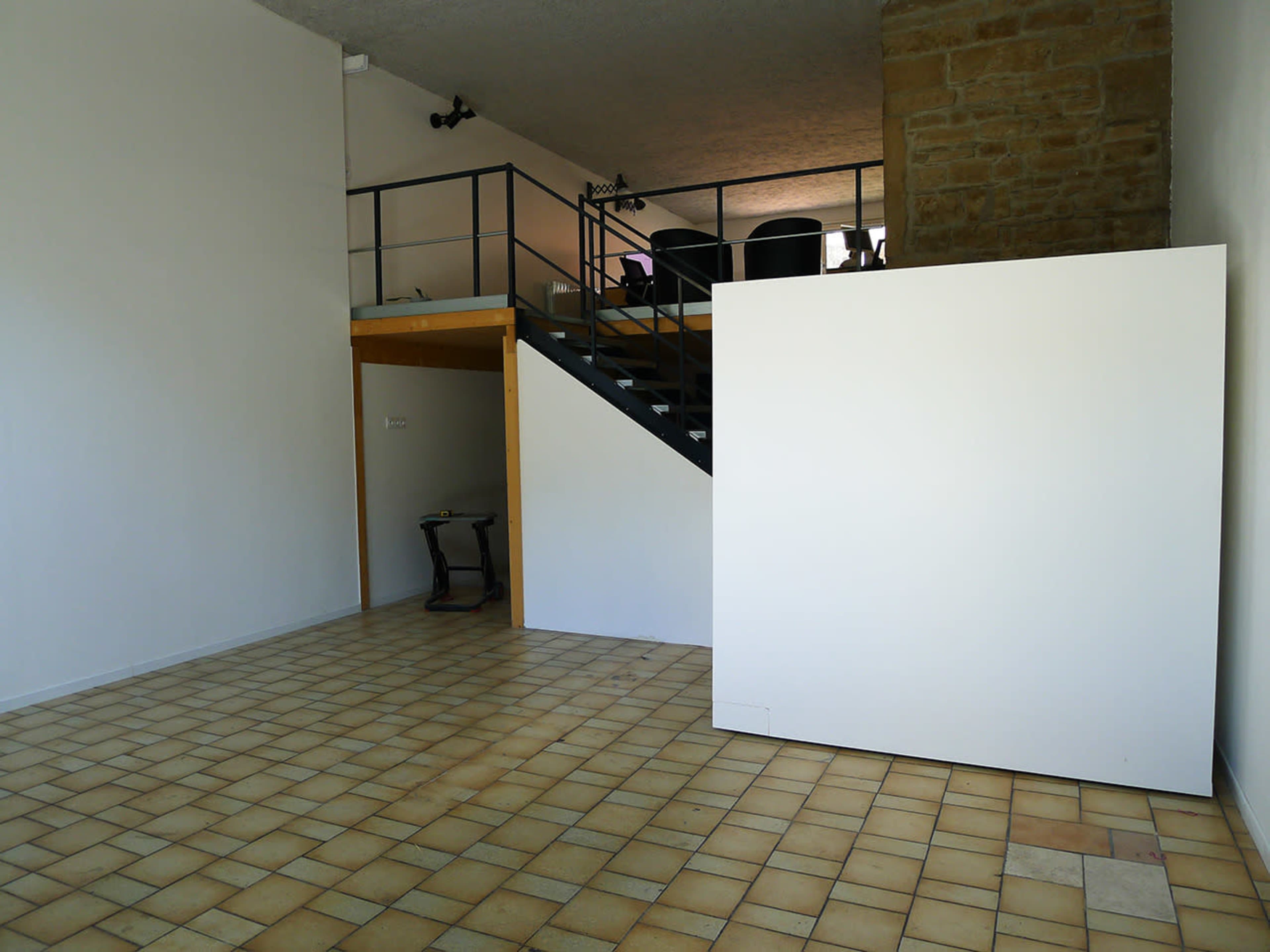 The image shows an empty interior space featuring tiled flooring, a staircase leading to a loft area, and a large white partition against one wall.