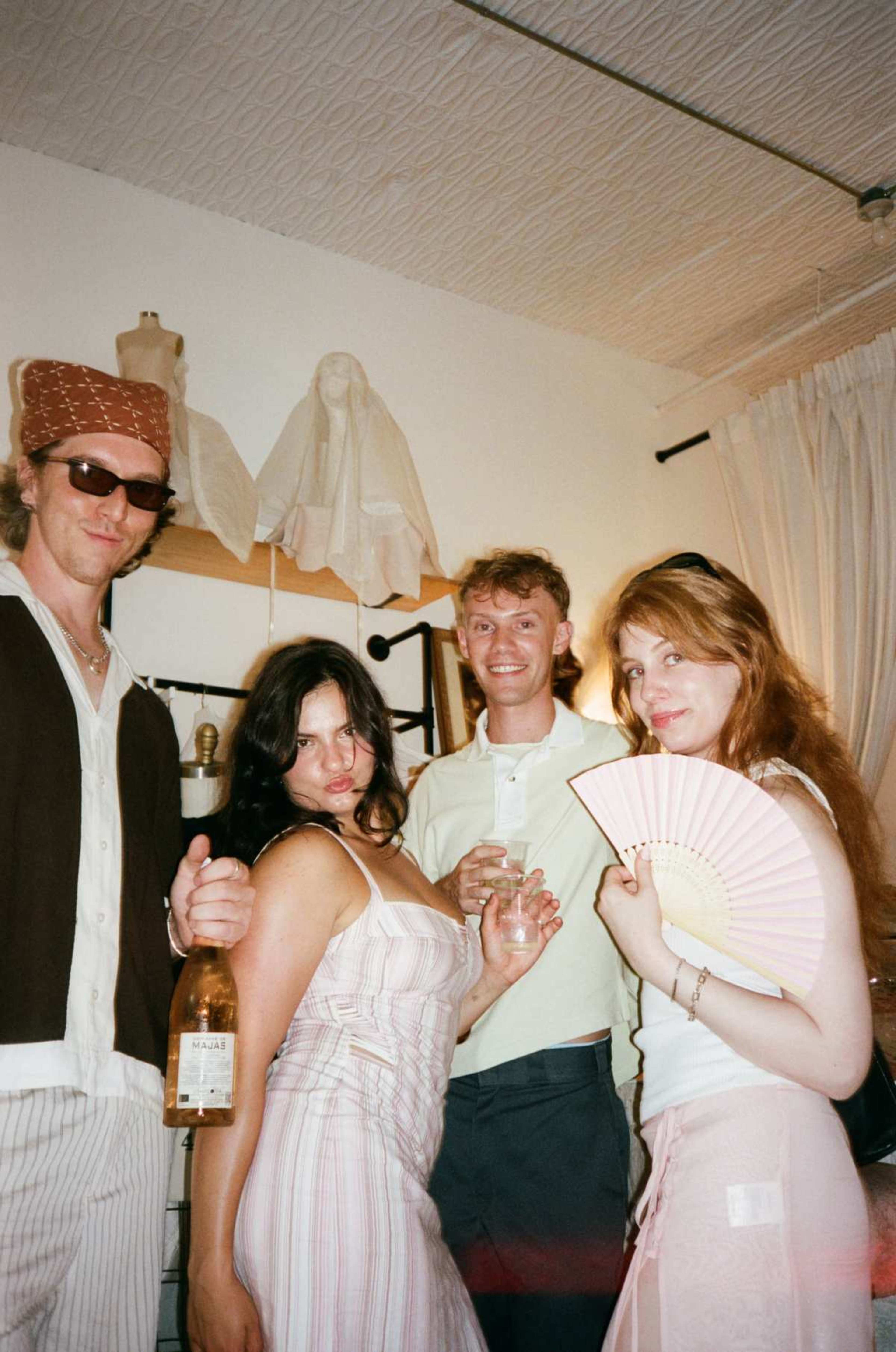 Four people pose together indoors, each holding a drink, with a fan held by one individual and various mannequins and fabric drapes in the background.
