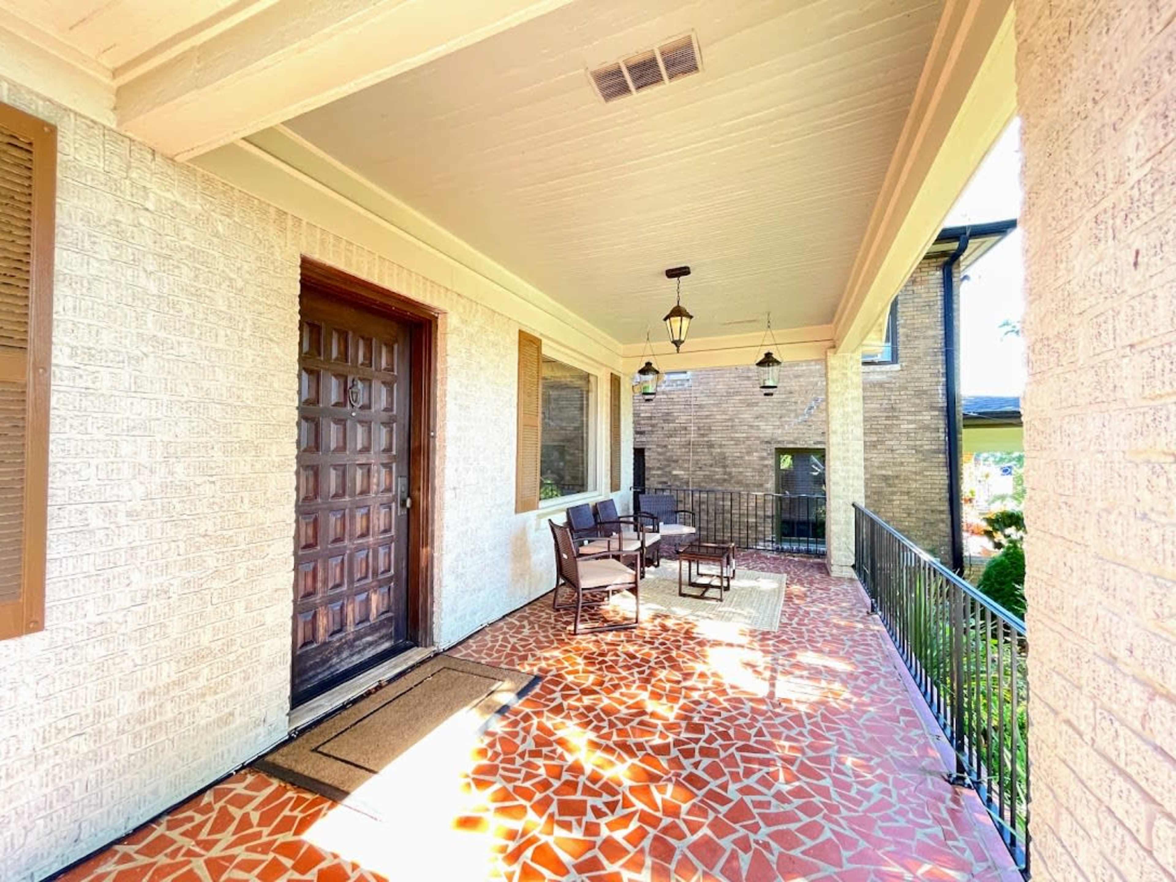 A covered porch with a wooden front door, a seating area with chairs and a table, and a patterned tile floor.