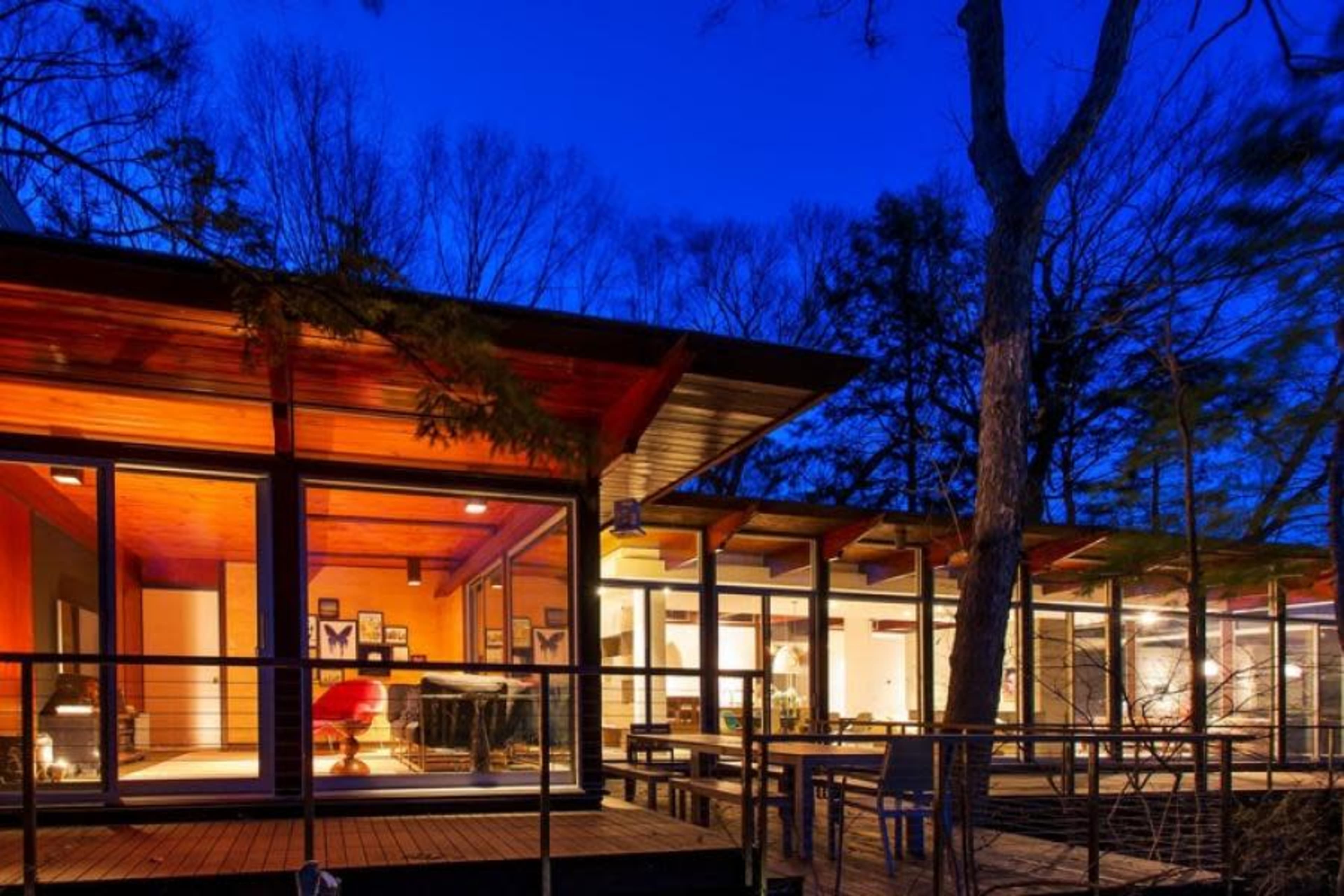 The image shows a modern house with large glass windows, illuminated interior spaces, and a deck surrounded by trees at dusk.