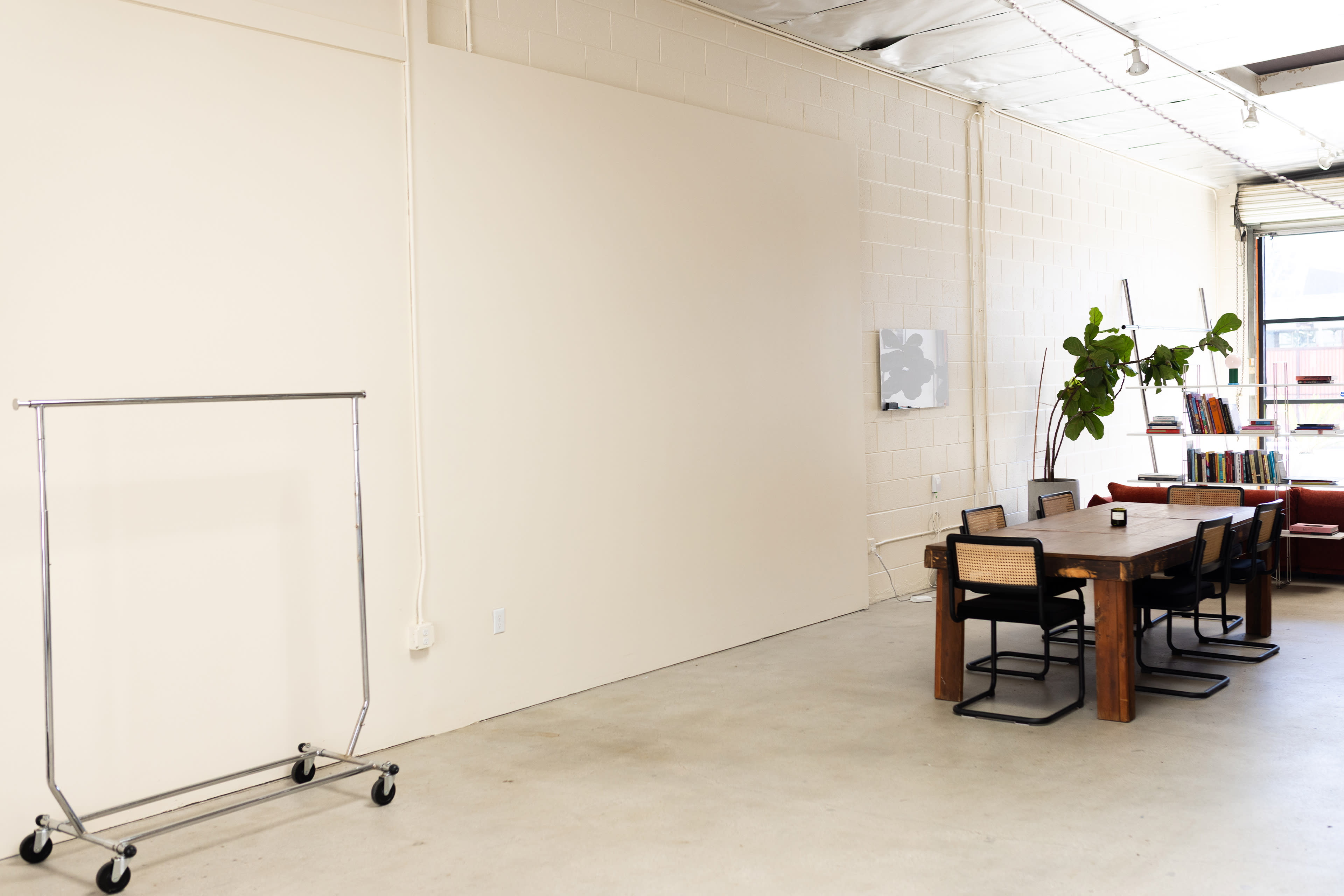 The image shows a spacious, minimalist interior with a dining table surrounded by chairs, a plant in the corner, a clothing rack on wheels, and a wall-mounted artwork.