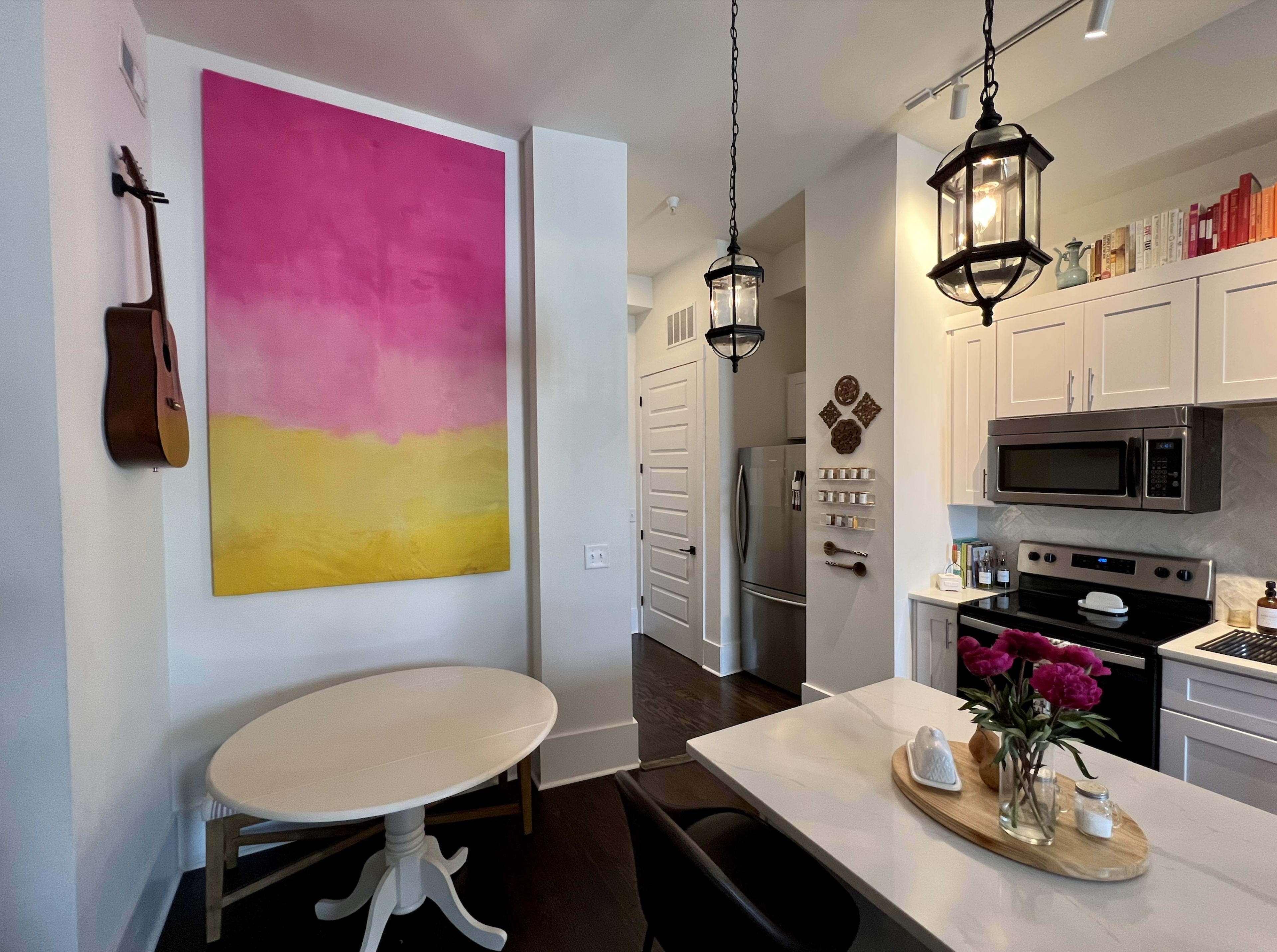 A modern kitchen features a large pink and yellow abstract painting on the wall, a round white dining table, and stainless steel appliances.
