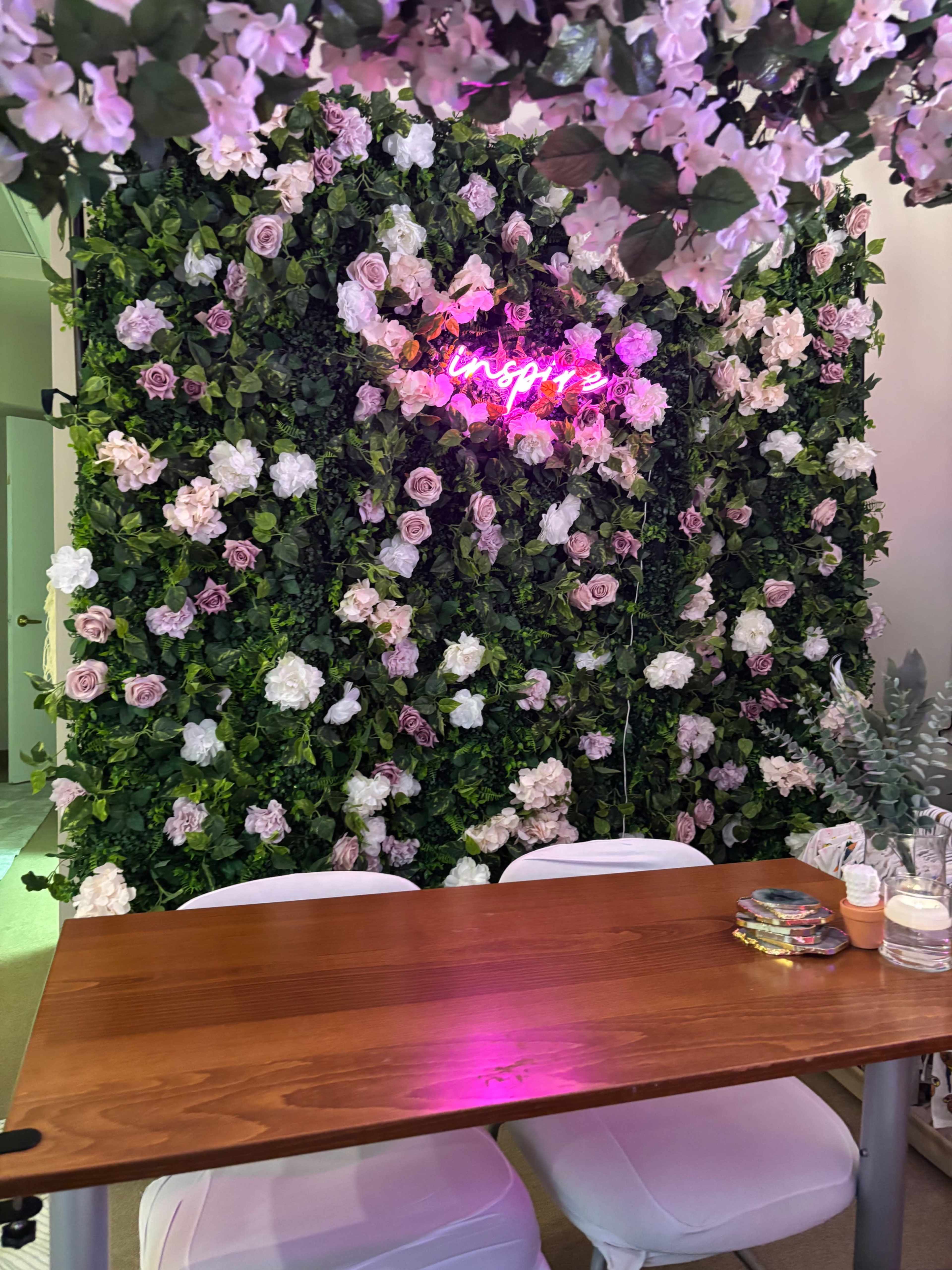 The image shows a decorative wall covered in artificial flowers, with a neon sign that reads "Inspired," set behind a wooden table with white chairs.