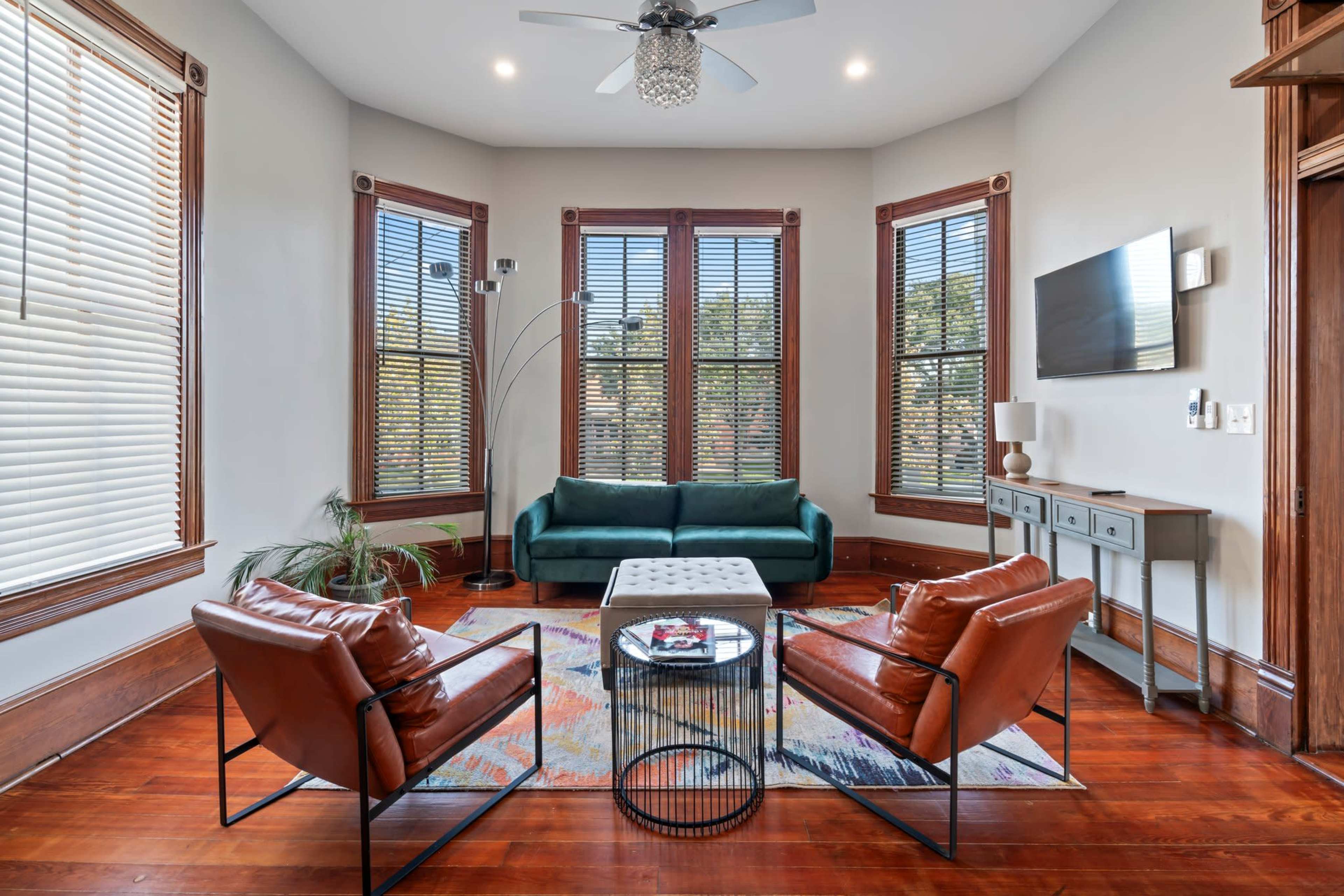 A living room features a green sofa, two brown chairs, a coffee table, and large windows with blinds.