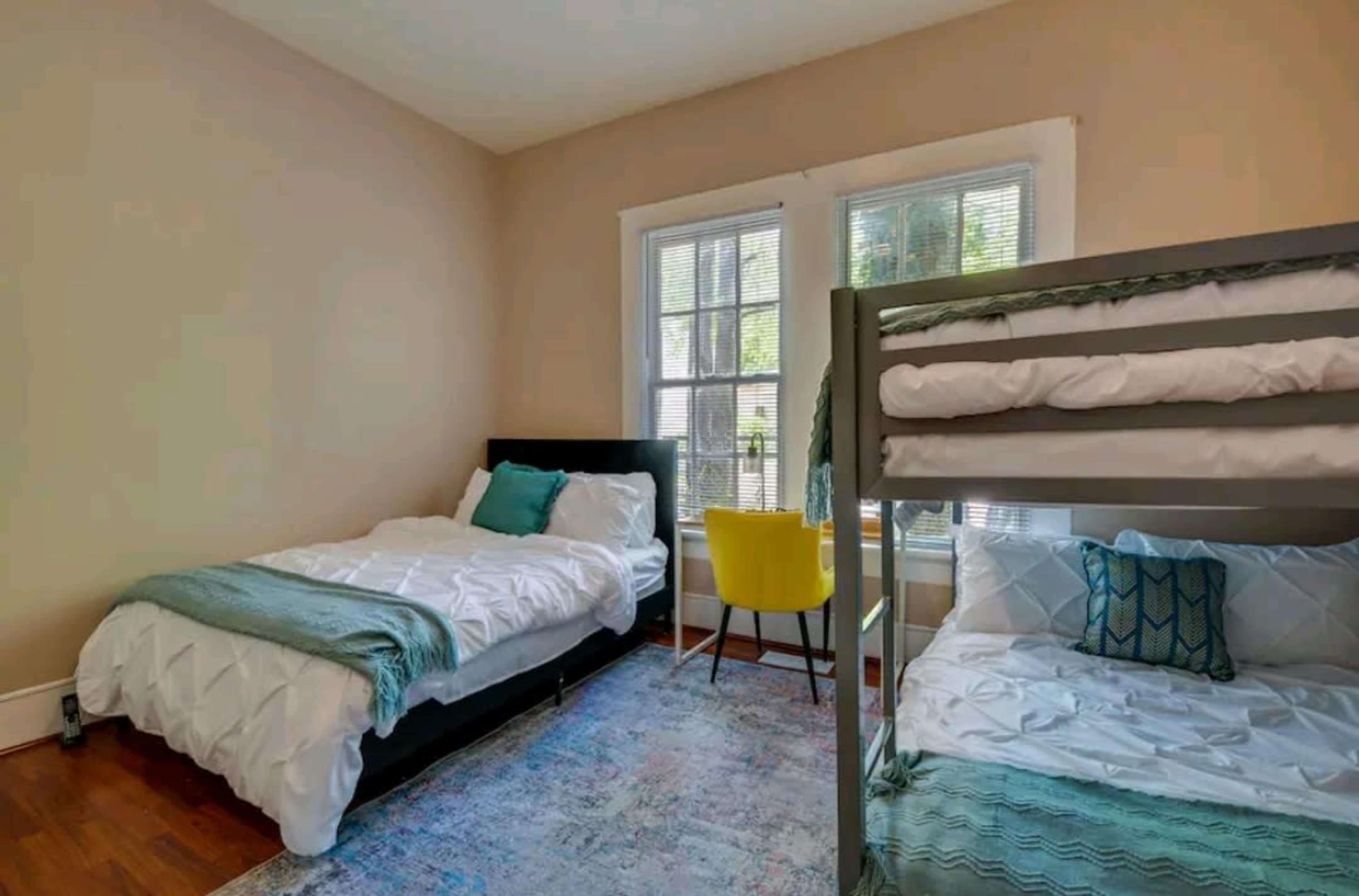 The image shows a bedroom with a double bed, a bunk bed, a small yellow chair, and two windows allowing natural light.
