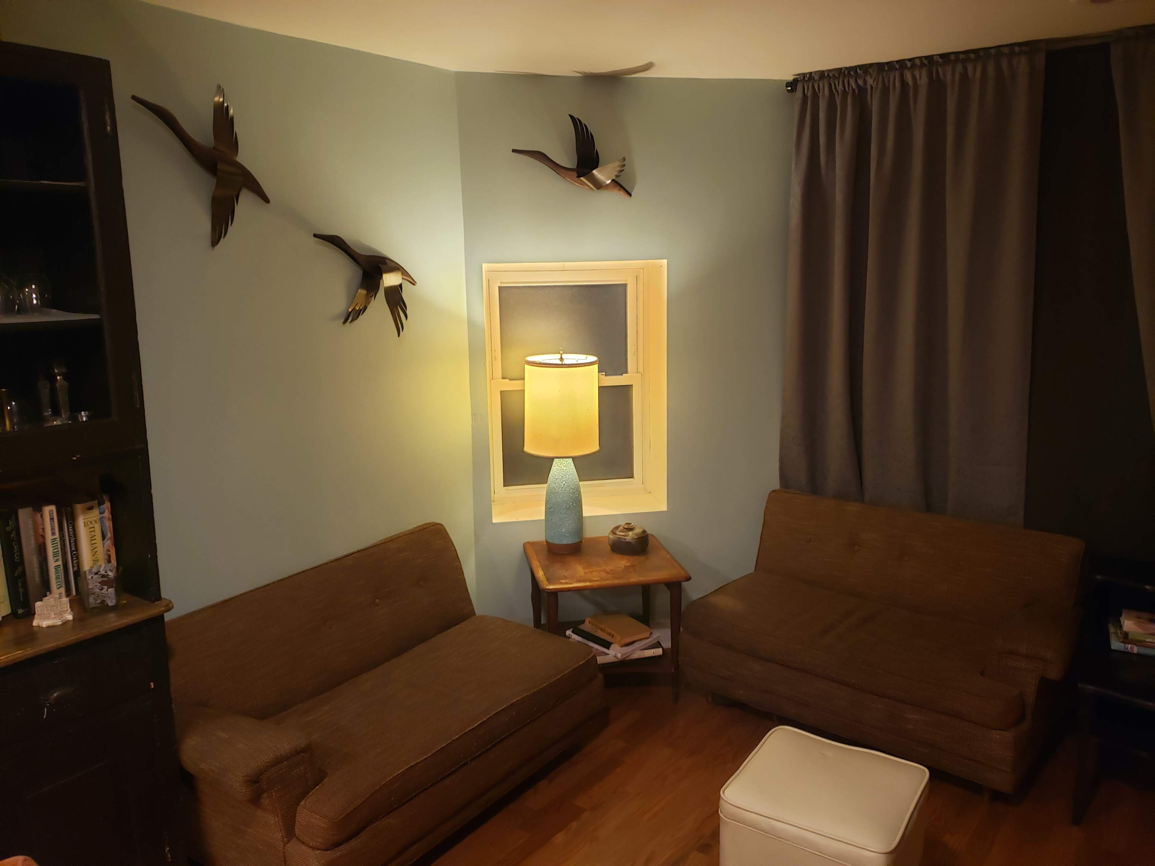 A cozy corner of a room with two brown couches, a small wooden table, a lamp with a blue base, and three decorative bird sculptures on the wall.
