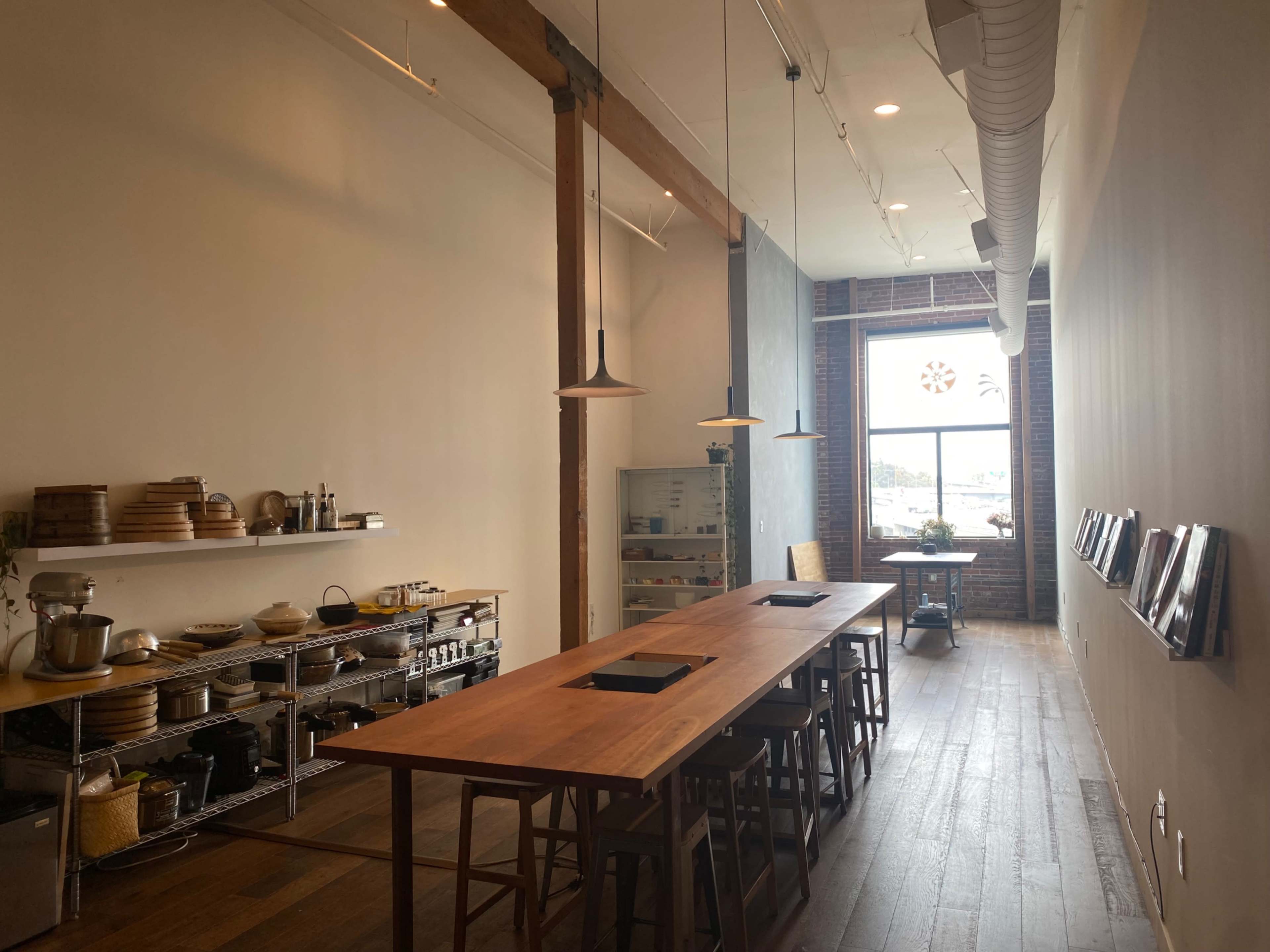 A spacious kitchen interior with a long wooden table, shelves of kitchenware, and a large window letting in natural light.