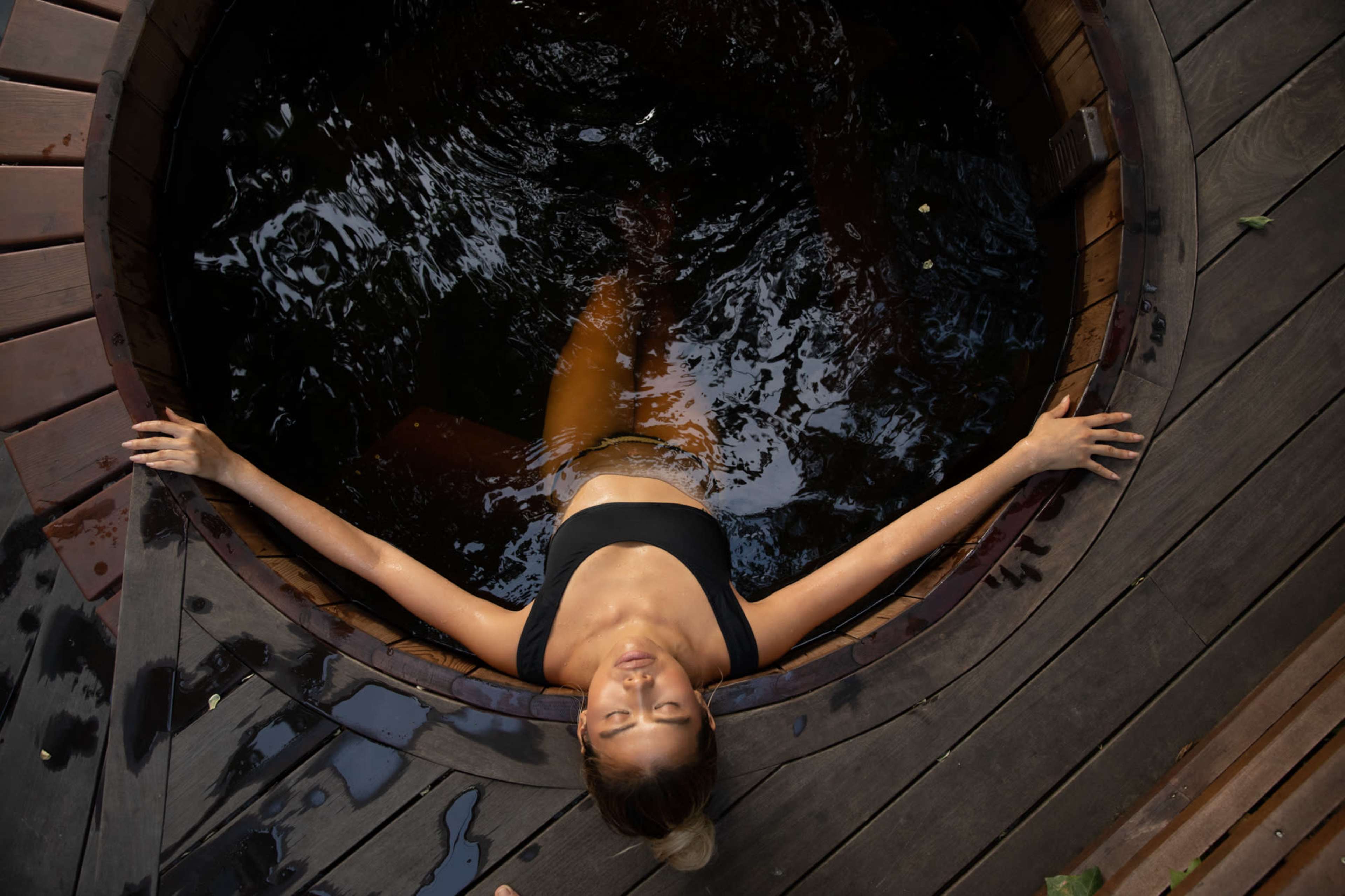 A woman relaxes in a wooden hot tub surrounded by dark water and wooden decking.