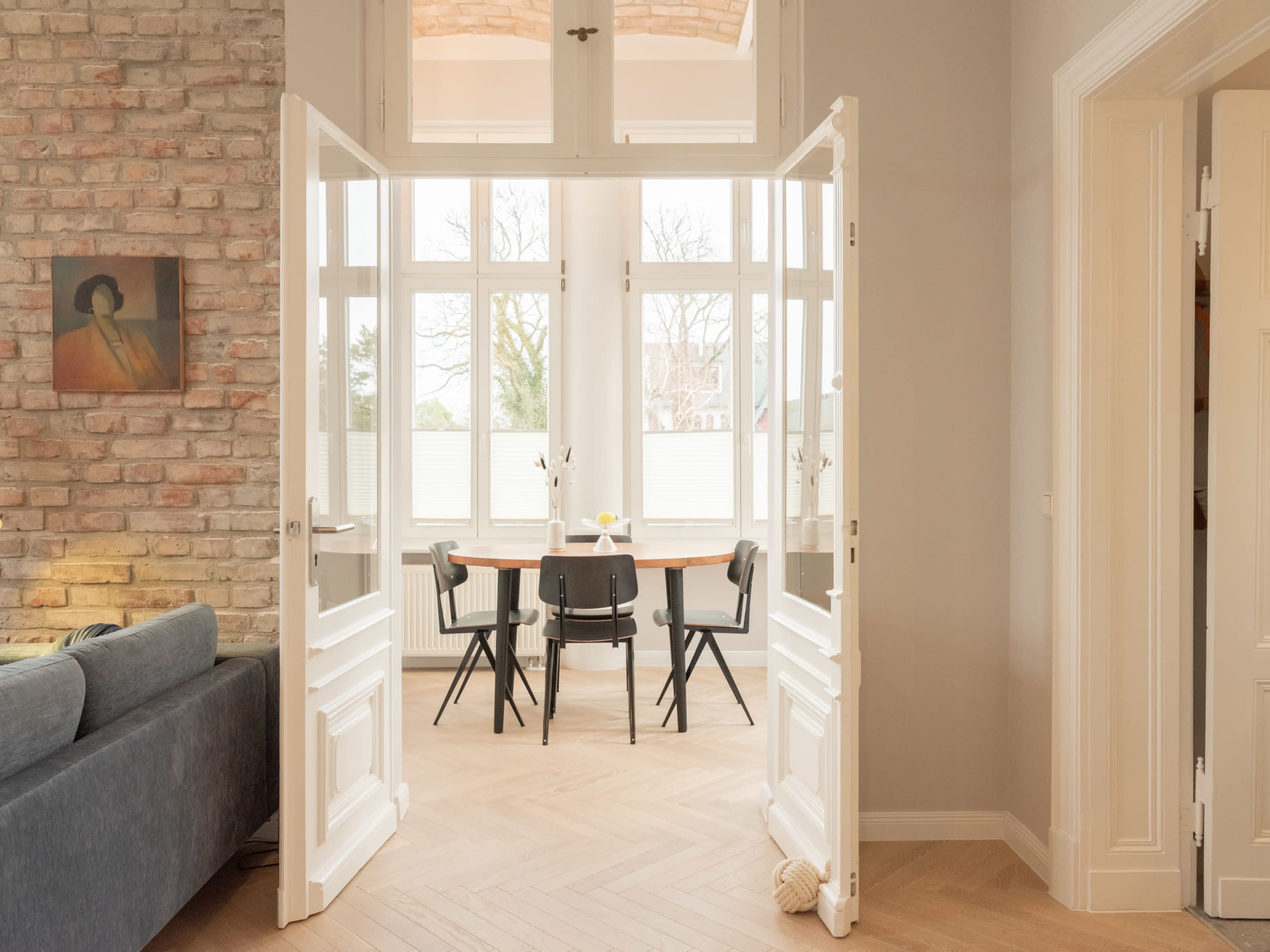 A set of white double doors opens to a dining area with a round table and four black chairs, surrounded by large windows that allow natural light to fill the space.