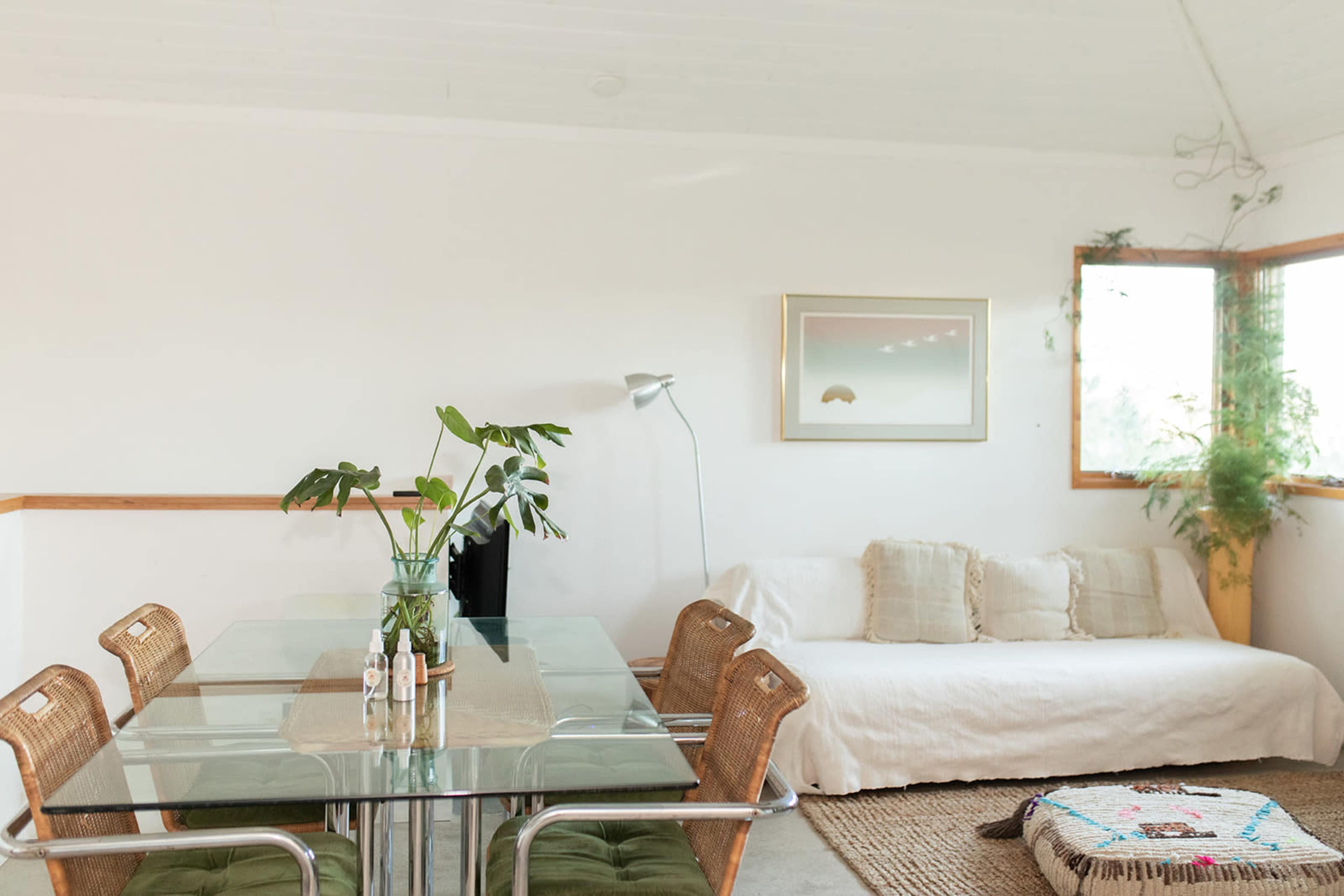 The image shows a bright, minimalist living space featuring a glass dining table with wicker chairs, a light-colored sofa, and a corner filled with plants.
