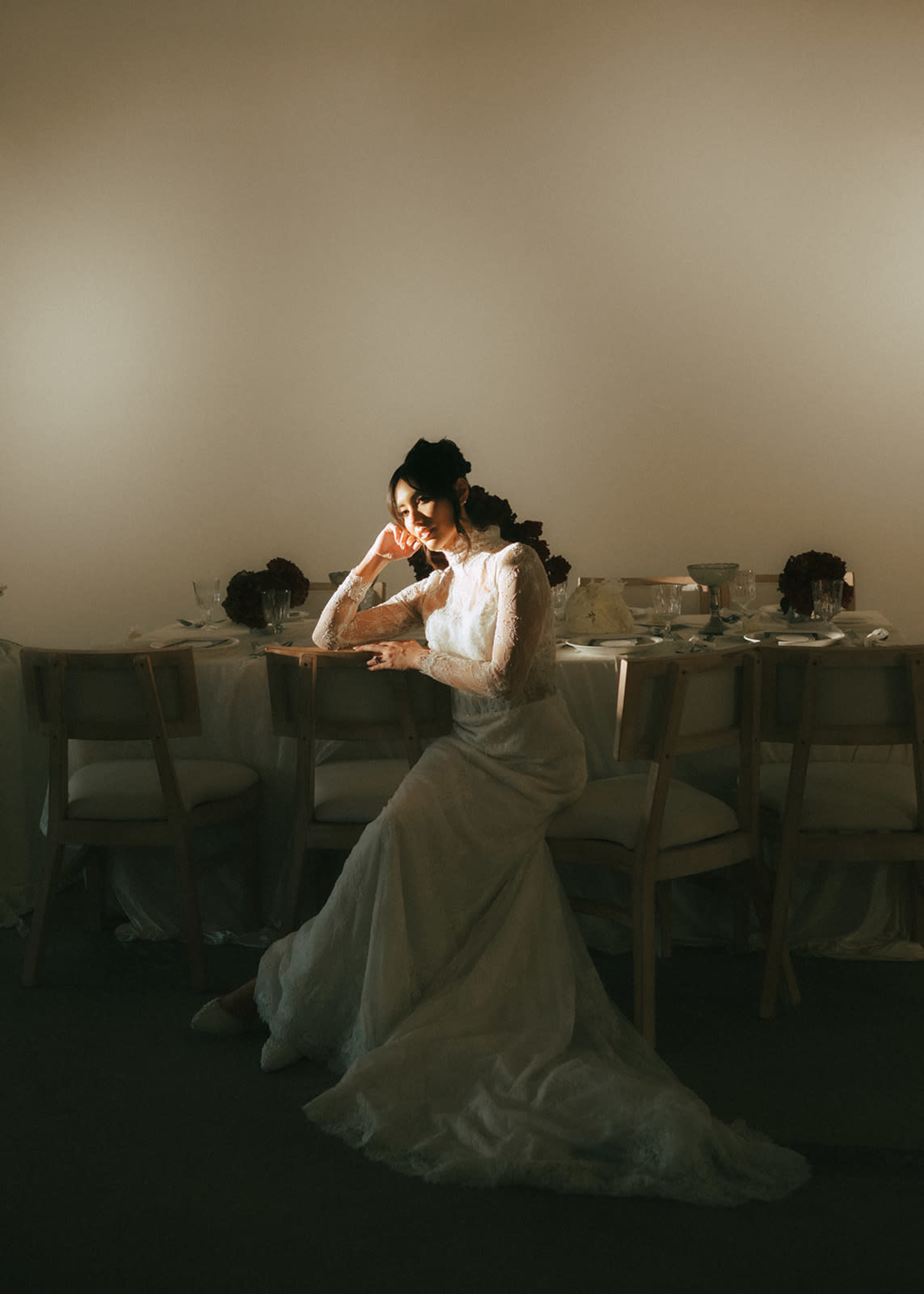 A woman in a wedding gown sits at a table, softly illuminated against a light background, with her head resting on her hand.