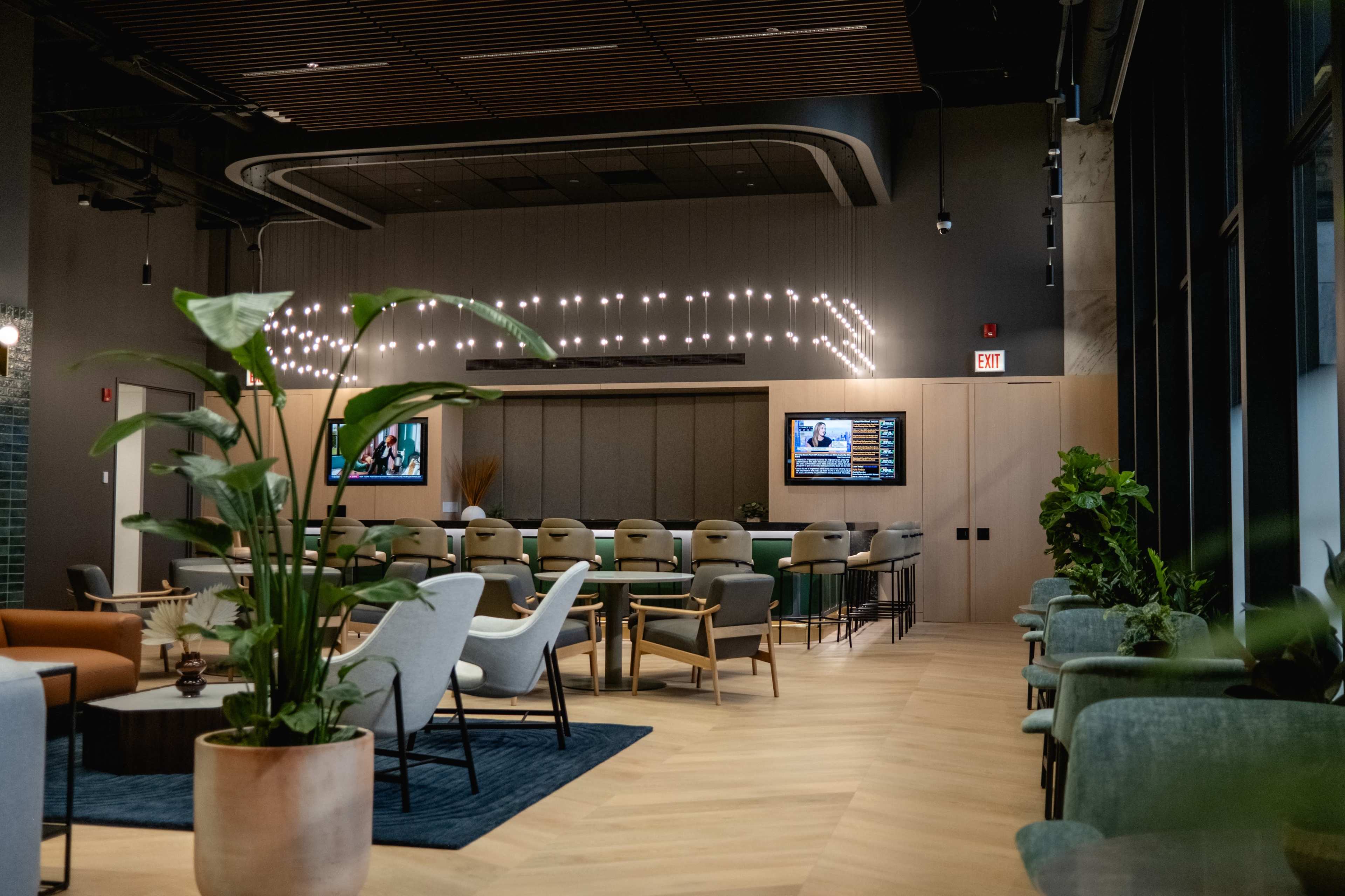 The image shows a modern lounge area with seating, tables, and screens displaying information, surrounded by greenery and wooden decor.