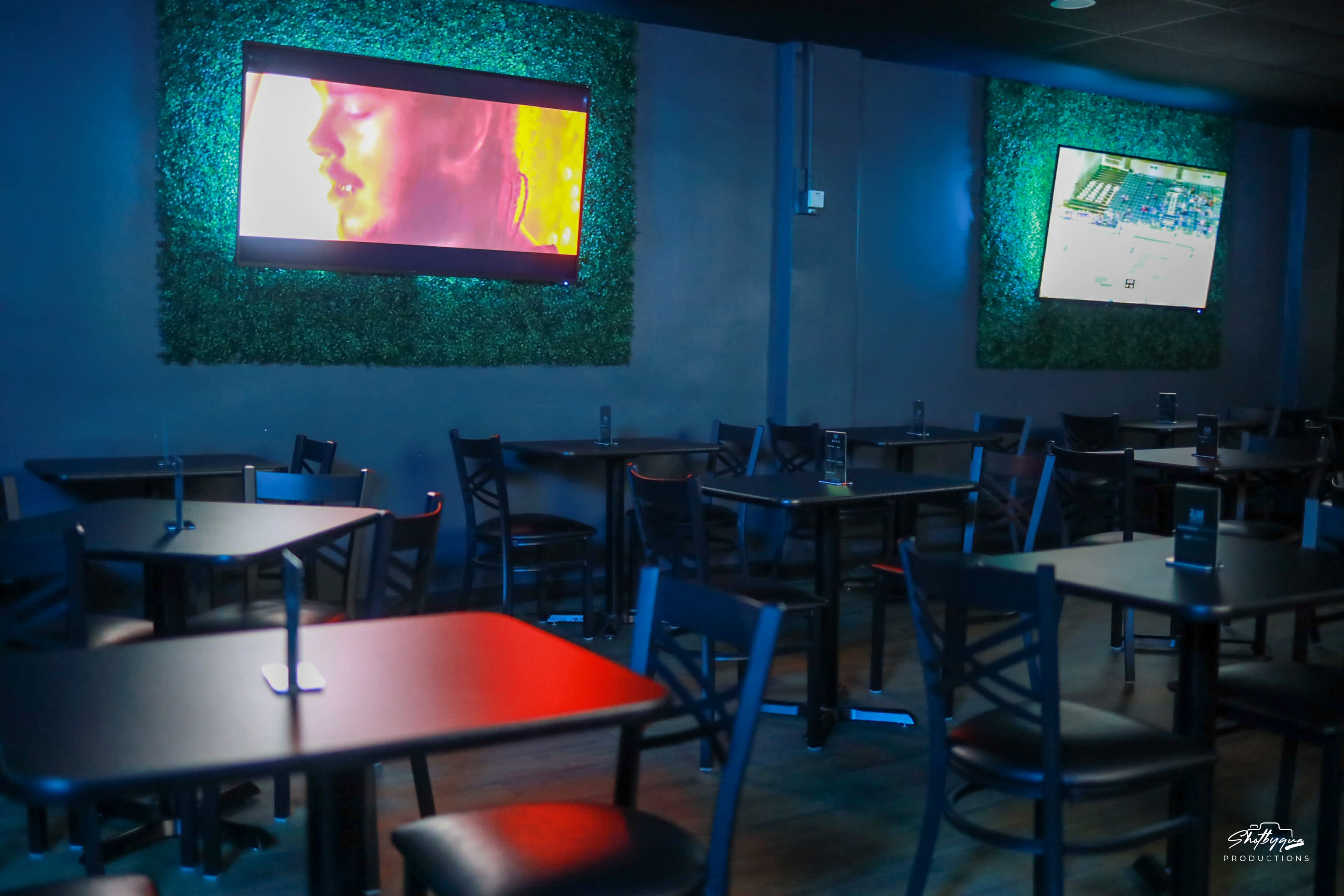 A dimly lit restaurant or bar with tables and chairs arranged around two wall-mounted screens displaying videos.