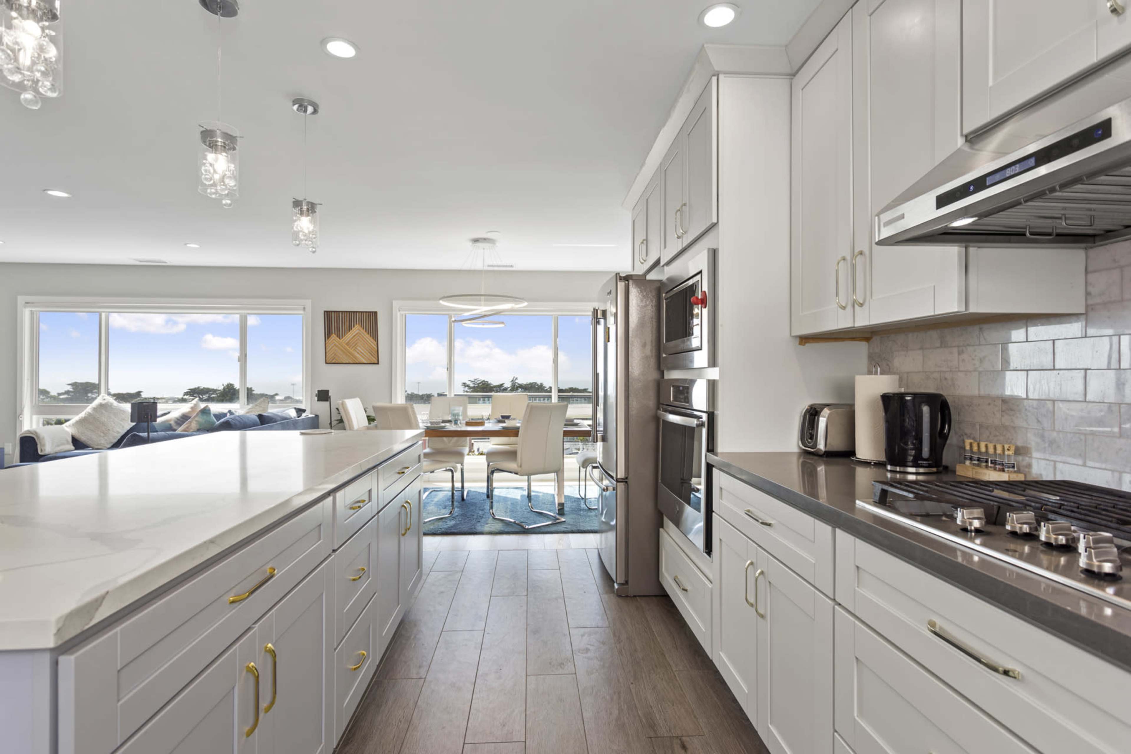 A modern kitchen features white cabinetry, stainless steel appliances, and a large island, with a view of a dining area and window overlooking a scenic landscape.