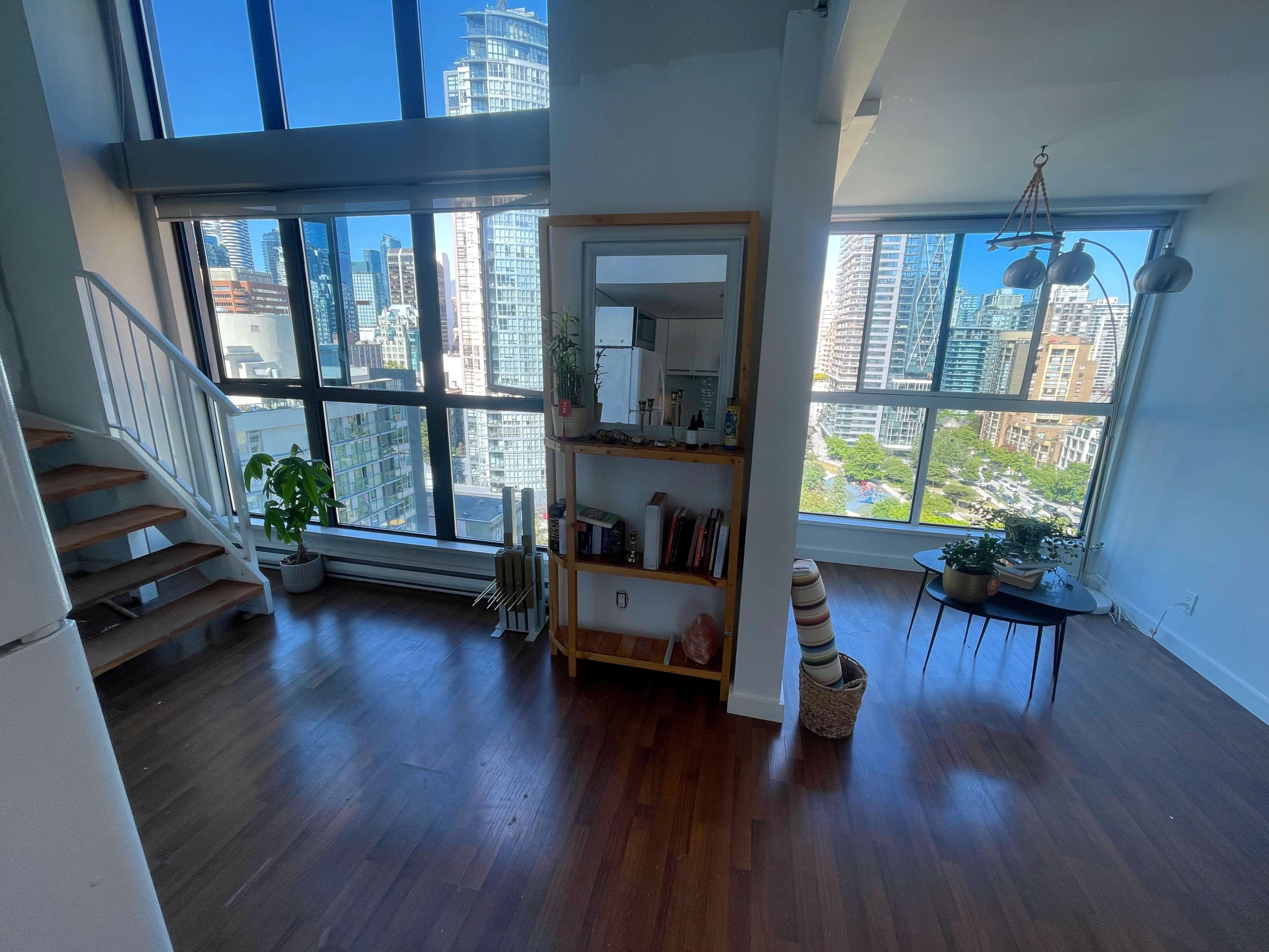 A modern, airy apartment with large windows overlooking a city skyline, featuring a wooden staircase and a small seating area with plants.