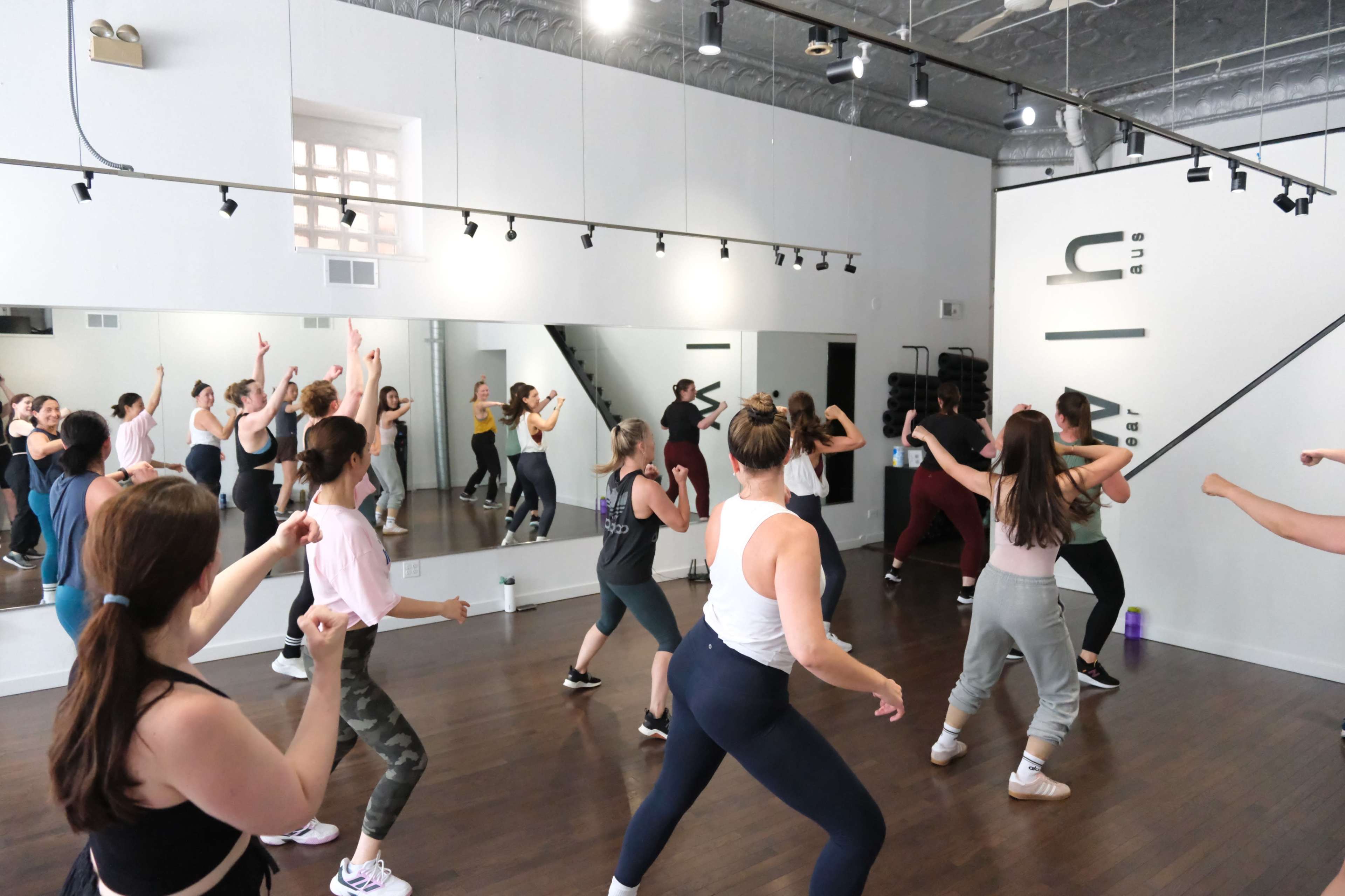 A group of people is participating in a dance class inside a spacious studio with mirrors and wooden floors.