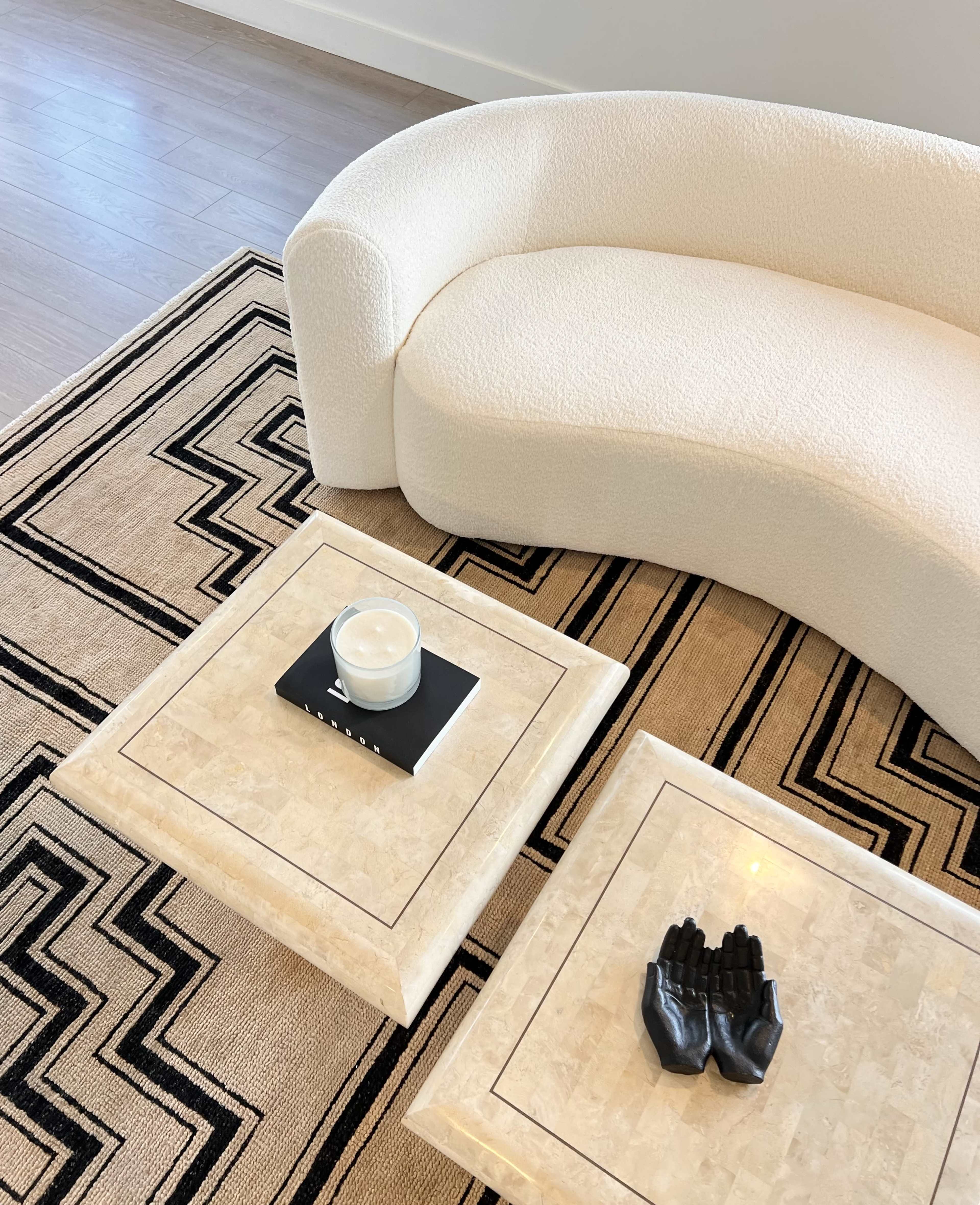 A textured white curved sofa placed above a geometric patterned rug, with two coffee tables made of light-colored stone, one holding a candle and the other displaying a black hand-shaped decor.