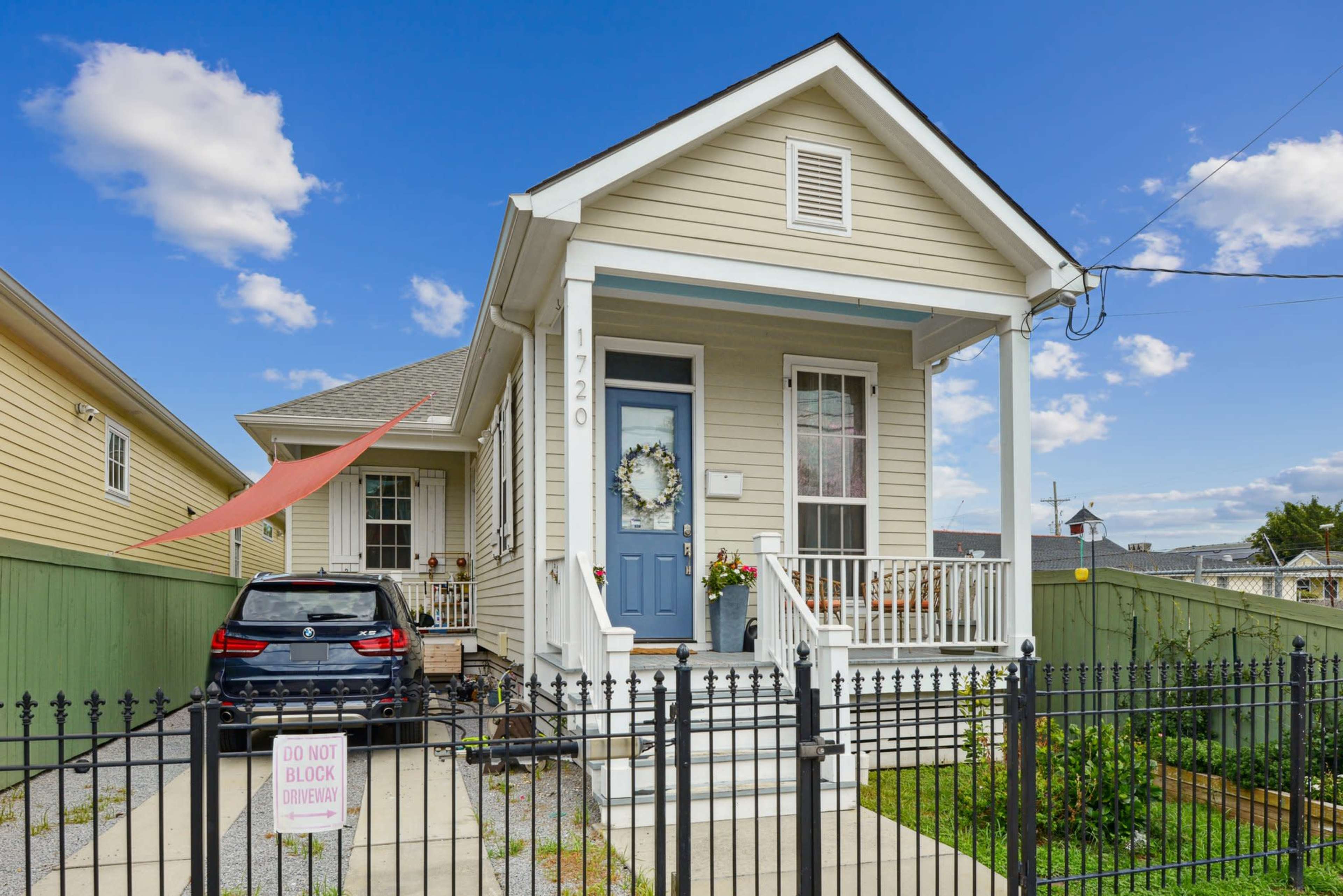 A light-colored house with a porch and a blue door is surrounded by a black fence, with a car parked in the driveway and a sign that reads "DO NOT BLOCK DRIVEWAY."