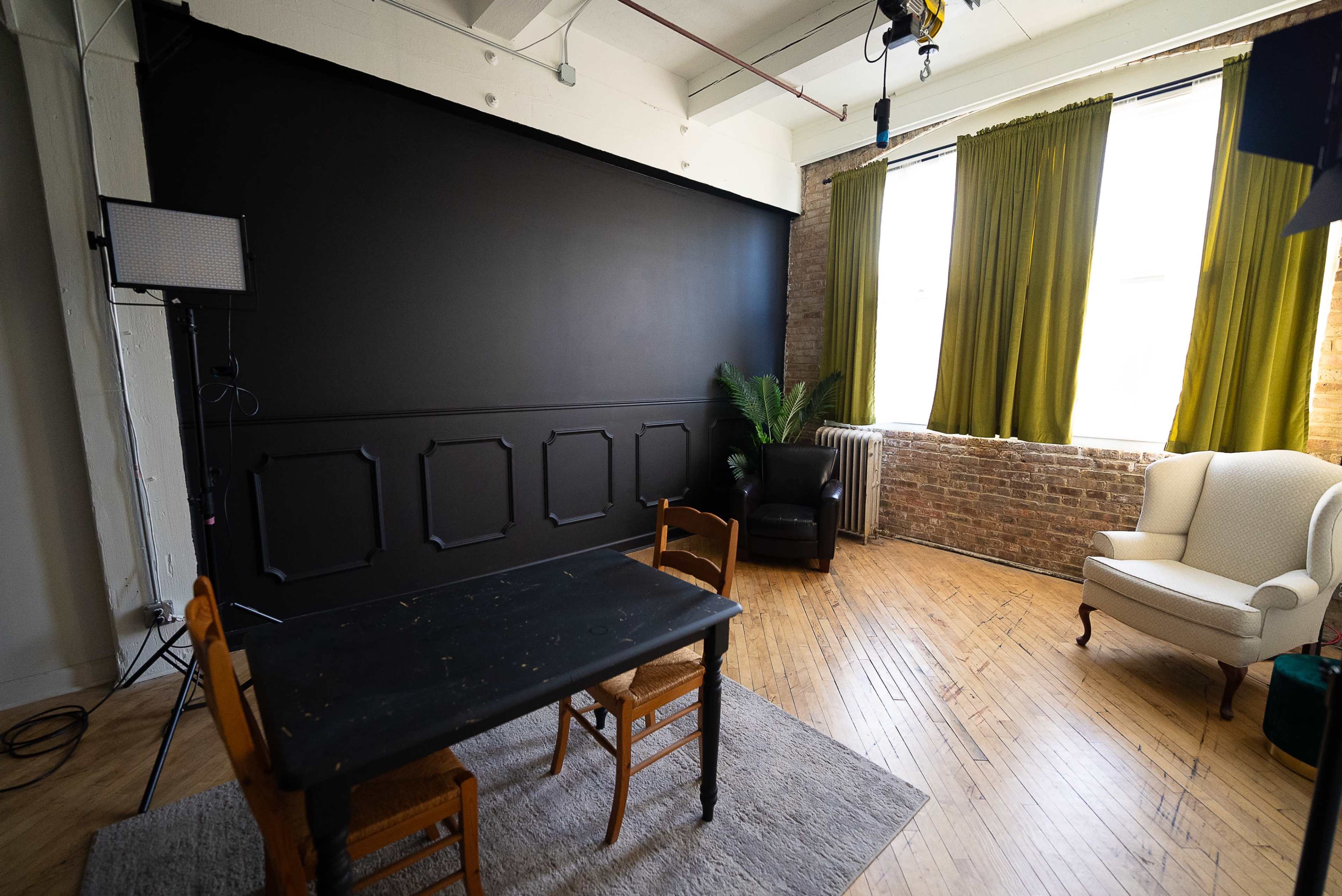 The image shows a well-lit studio space featuring a black backdrop, a wooden table with two chairs, and a cozy seating area with a green curtain window.