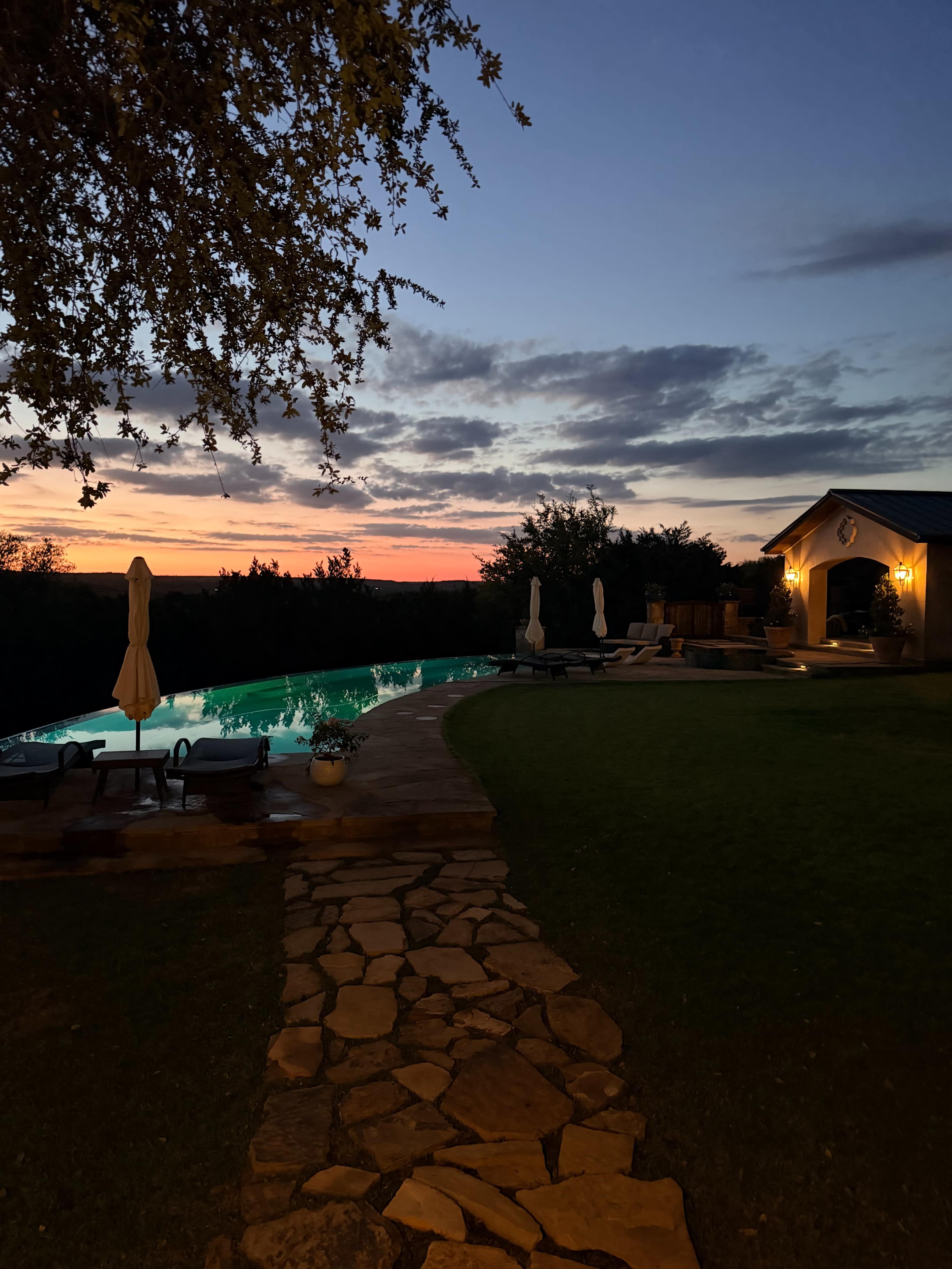 A stone pathway leads to a poolside setting with lounge chairs and a building, under a colorful sunset sky.