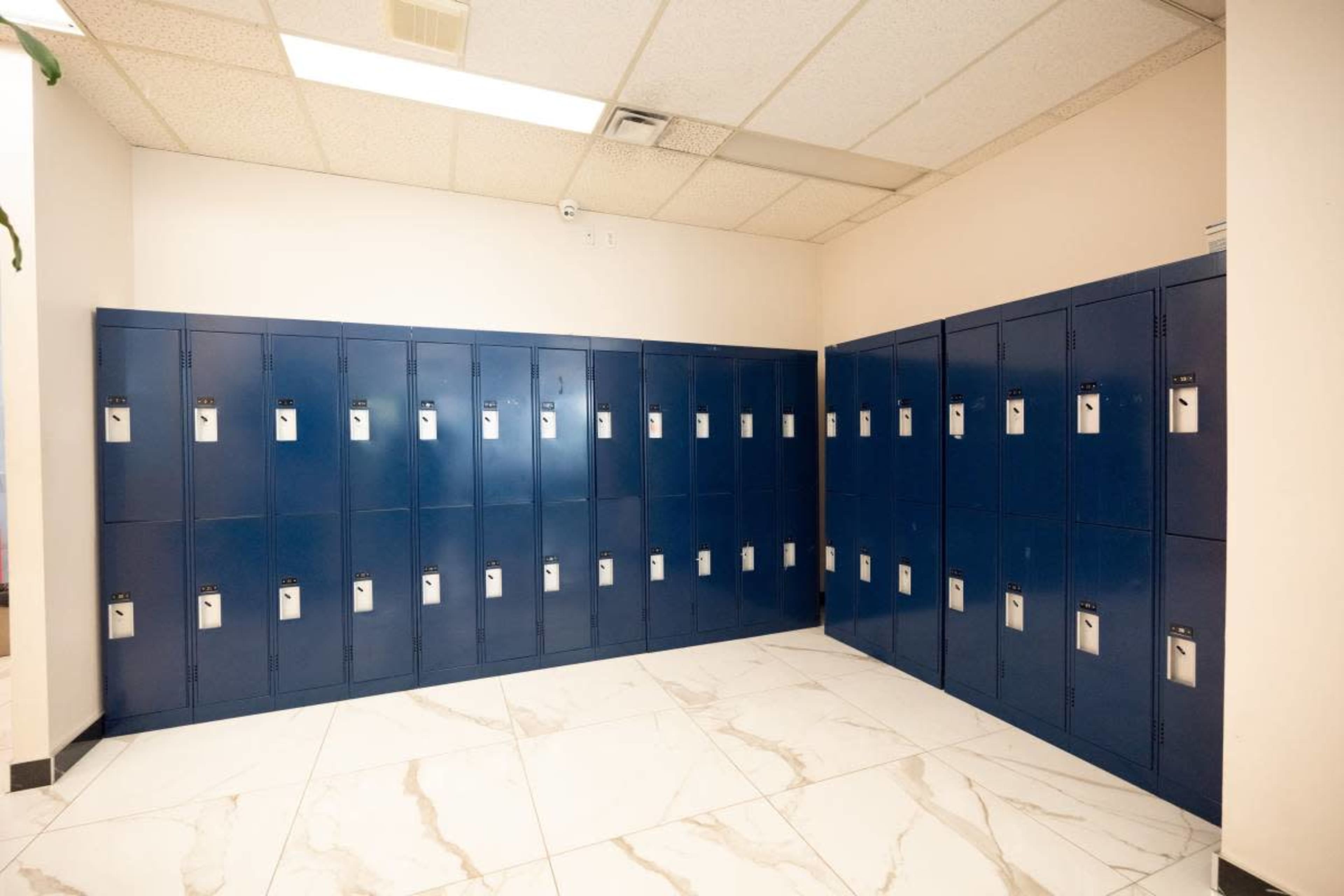 The image shows a row of blue lockers lined against a wall in a corridor with tiled flooring.