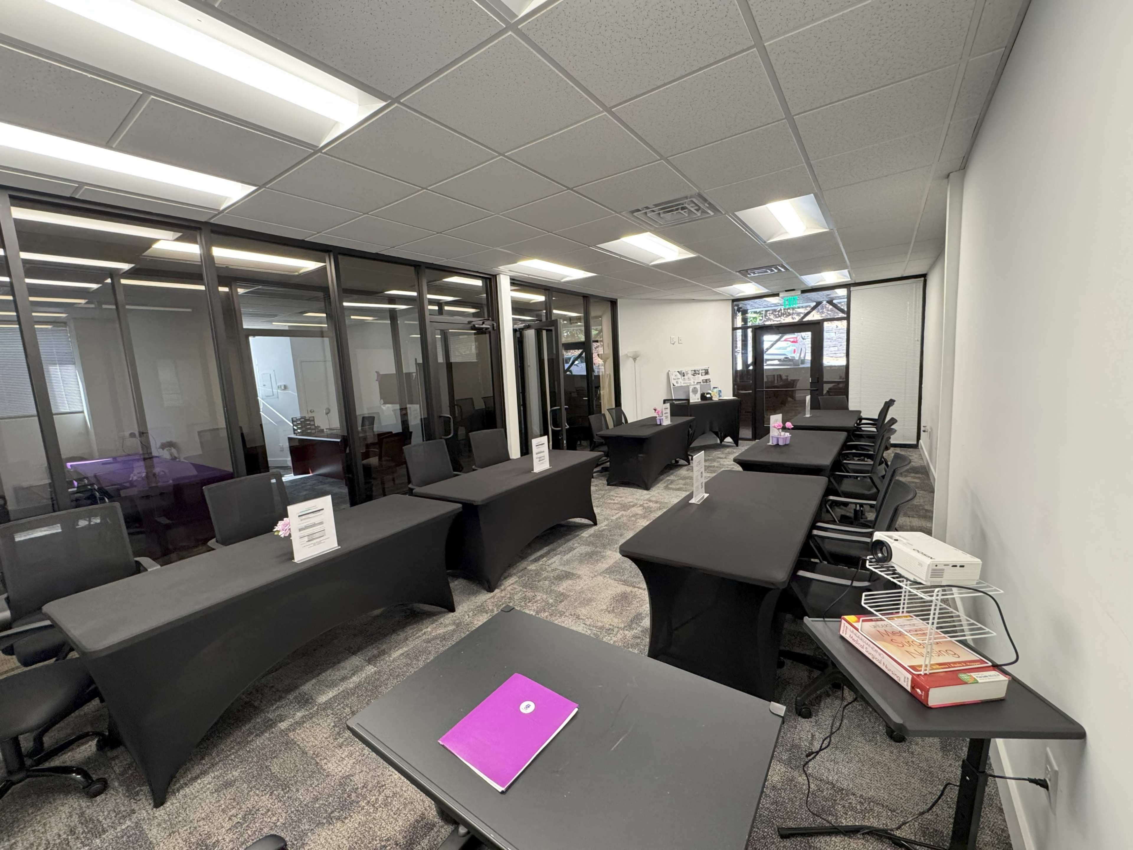 The image shows a modern office space with several black desks arranged in a semi-open layout, surrounded by glass-enclosed meeting rooms.
