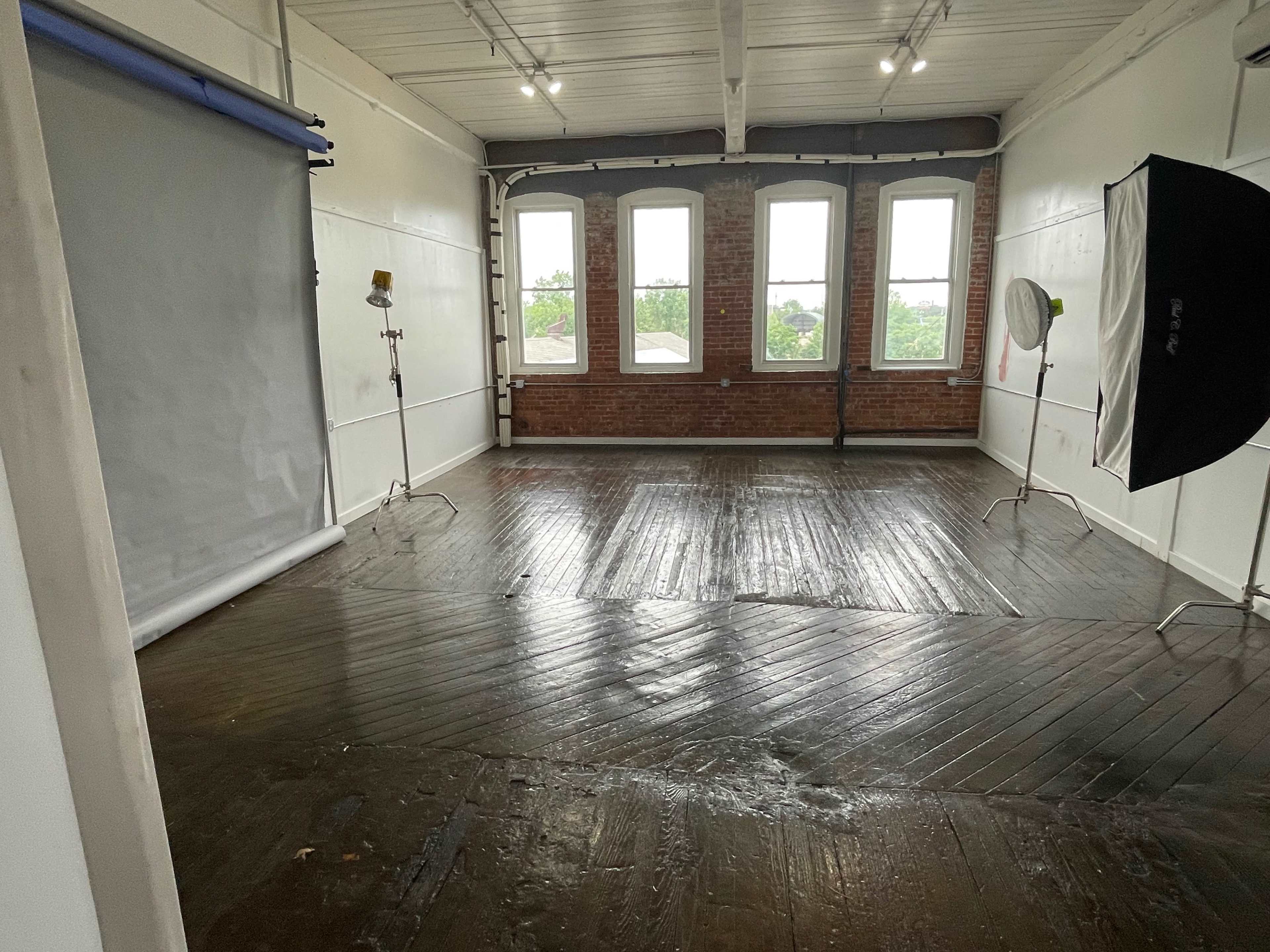 A photography studio with wooden floors, large windows, and lighting equipment set up on either side of a backdrop.