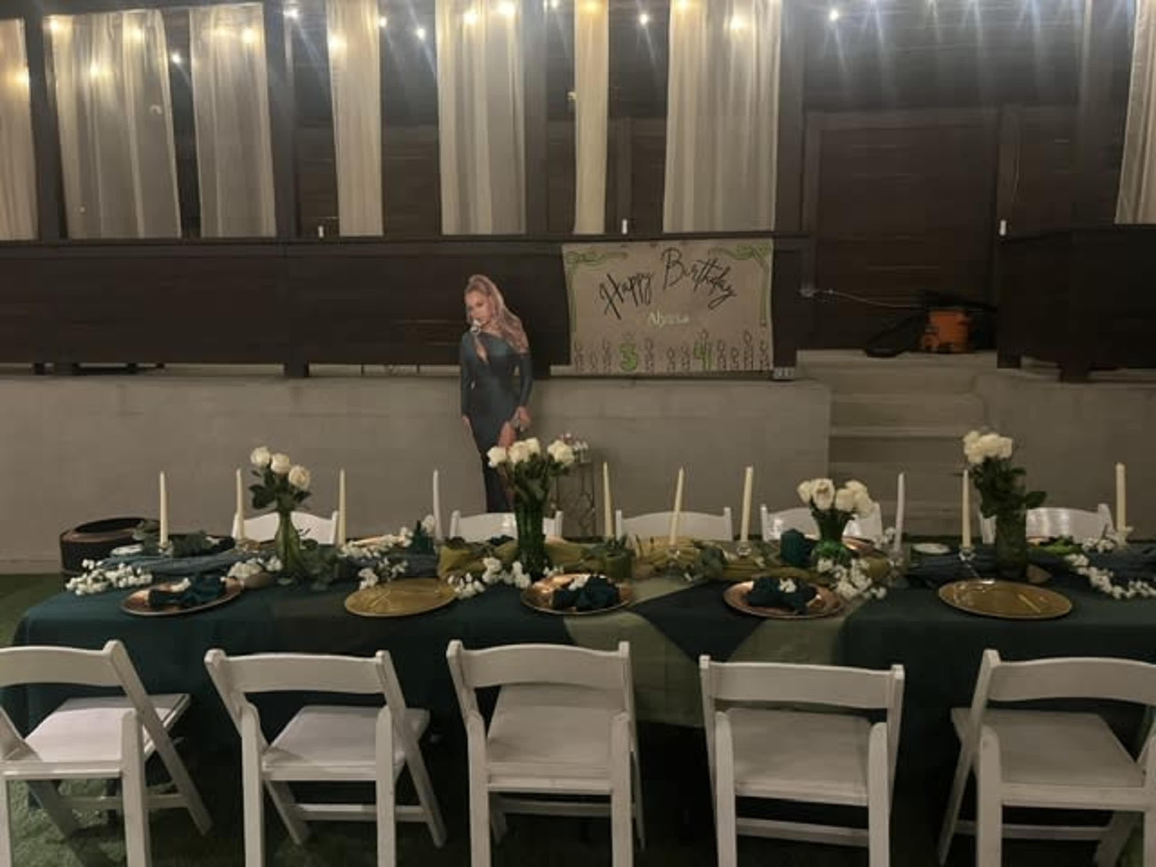 A long dining table set with plates, candles, and decorative flowers is arranged in a festive indoor space, with a birthday sign and a life-sized figure in the background.