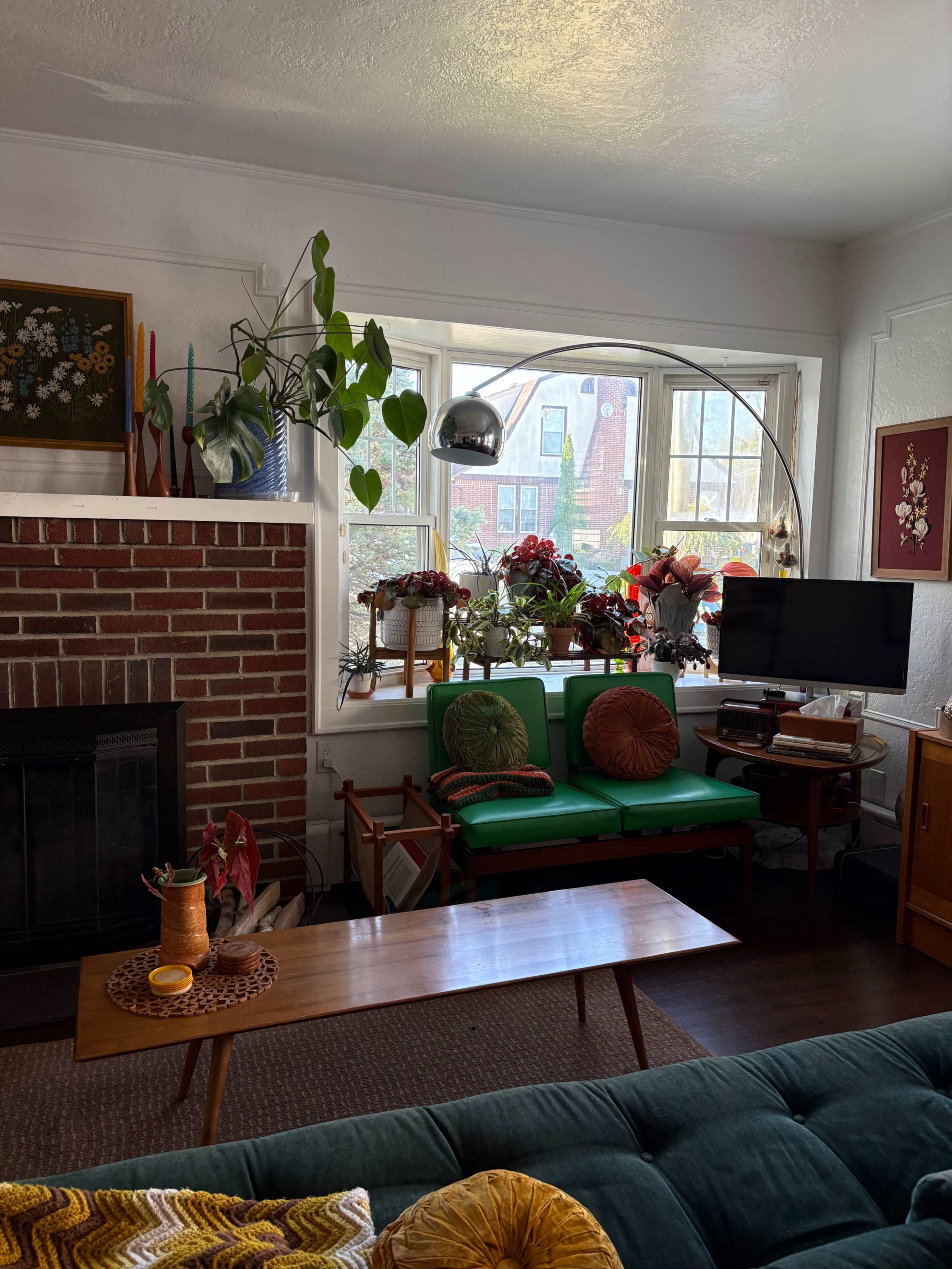 The living room features a green sofa, a wooden coffee table, a brick fireplace, and a large window adorned with plants and colorful flower arrangements.
