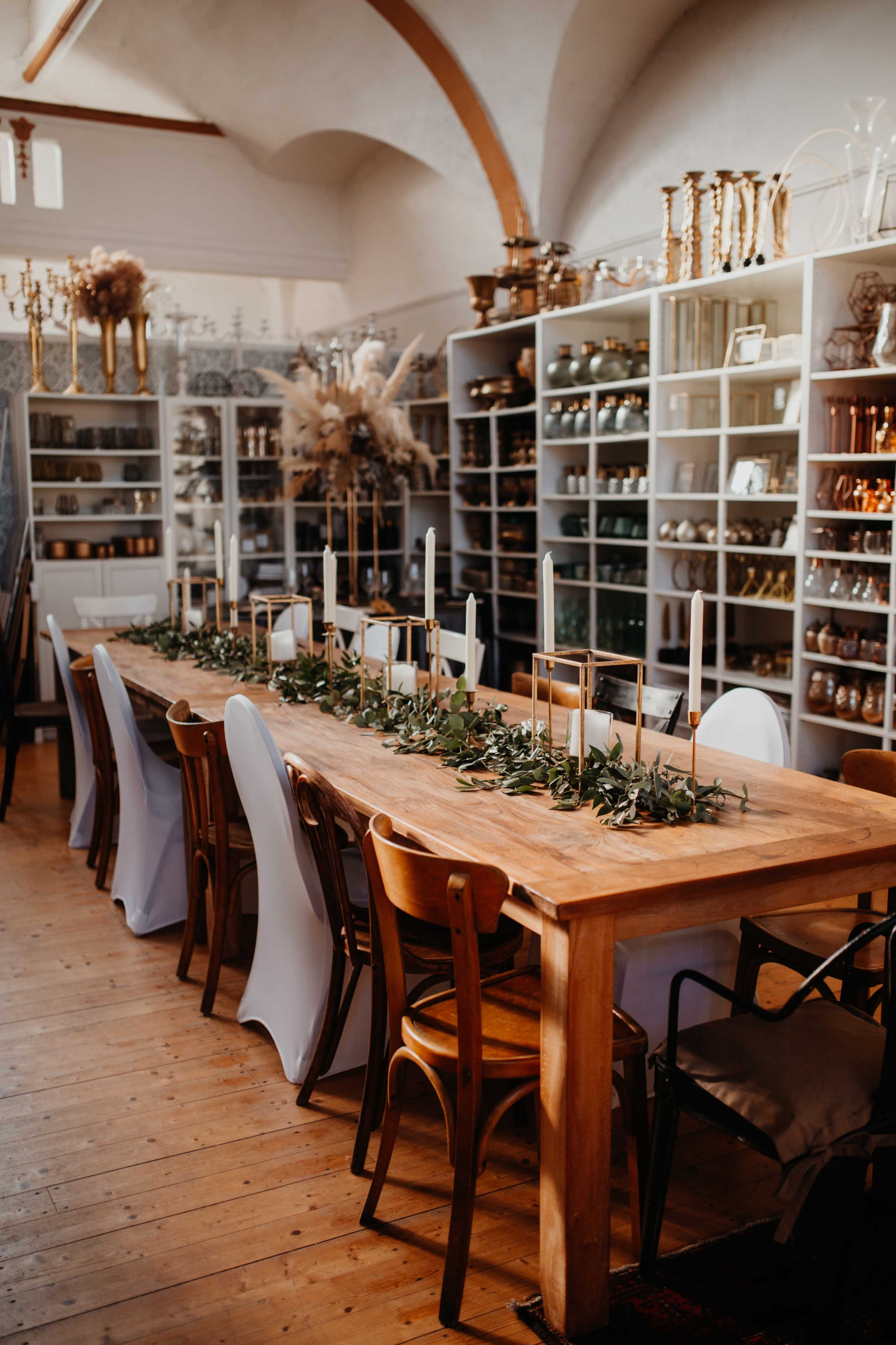 A long wooden dining table is set with candles and greenery, surrounded by chairs and display shelves filled with glass and metal decor.