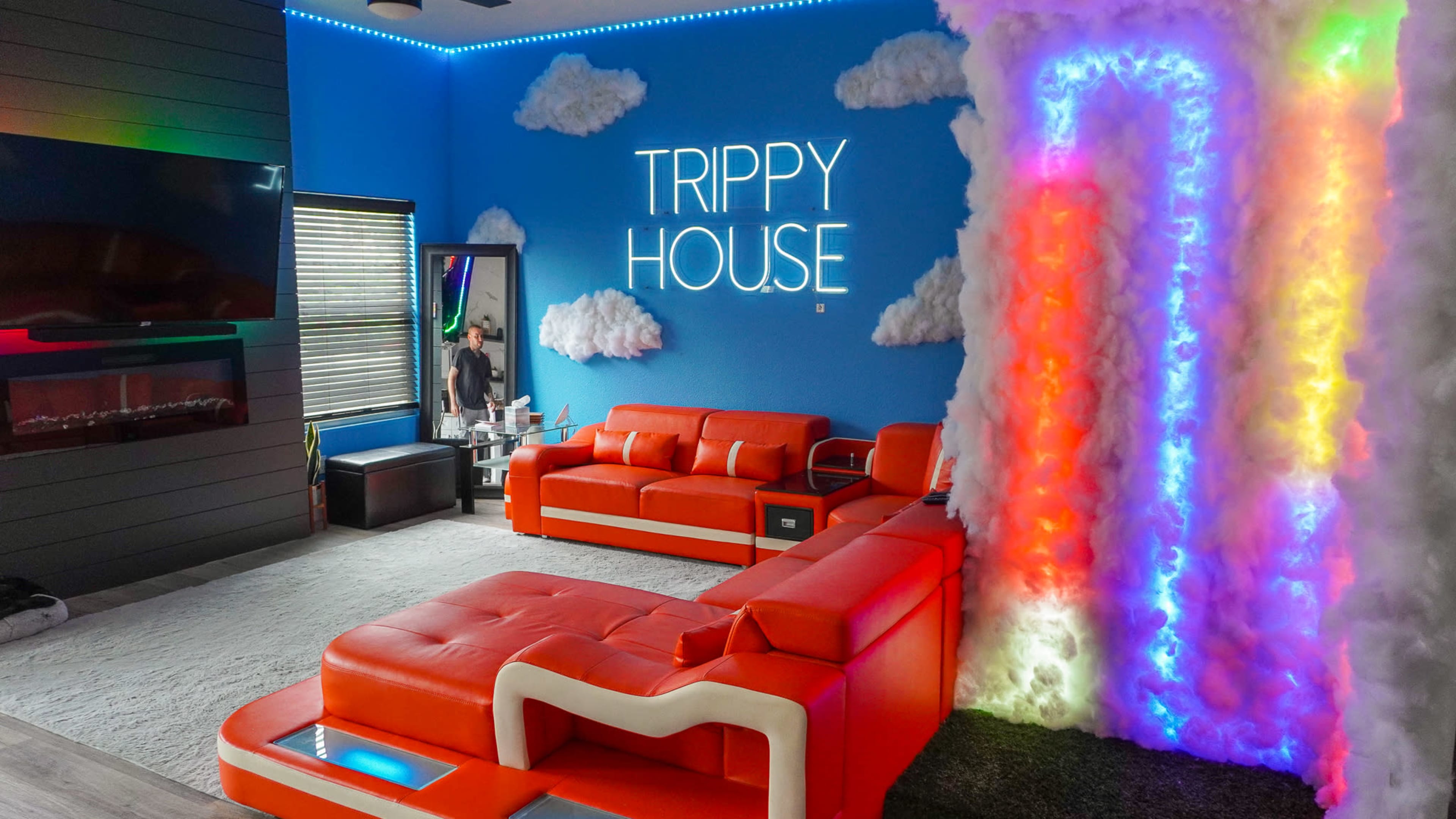 The room features bright orange furniture, a colorful cloud structure illuminated by neon lights, and a sign that reads "TRIPPY HOUSE" on a blue wall.